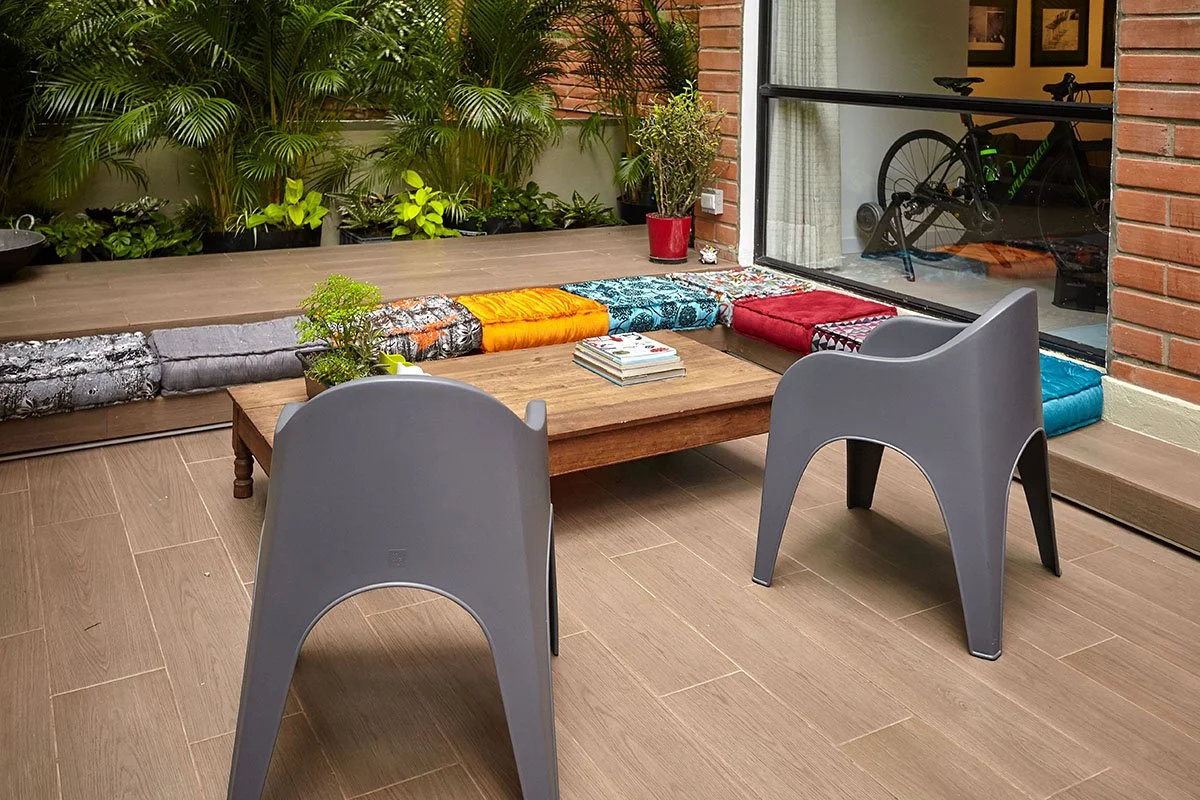 Sala con dos sillas grises, mesa de madera, plantas en la esquina, cojines de colores sobre el piso, con ventana y bicicleta en el interior.