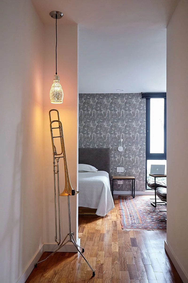Interior de una habitación con pared de ladrillos, cama con sábanas blancas, mesa de noche, alfombra colorida, escritorio con portátil, lámpara y trombón apoyados en la pared