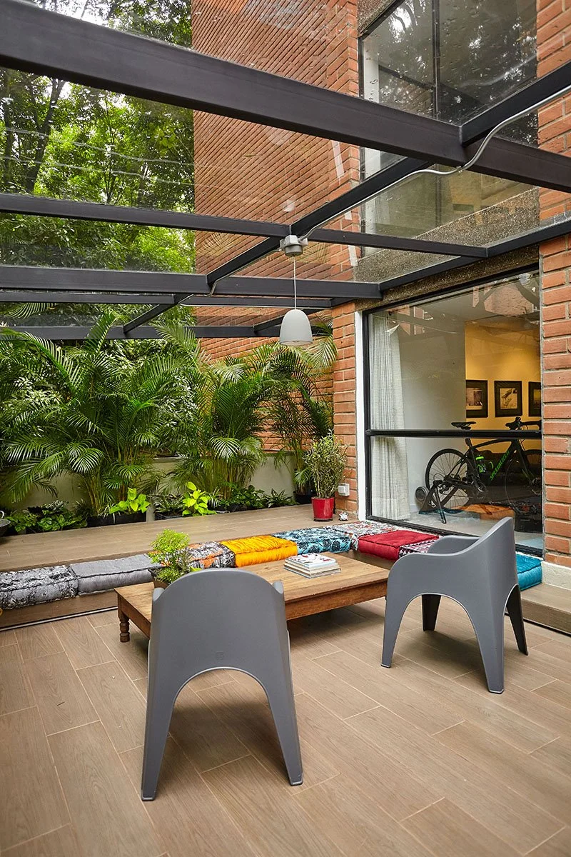 Un espacio de terraza con paredes de ladrillo, con un techo de vidrio y estructuras metálicas negras. Hay plantas verdes en la parte trasera, una mesa de madera con cojines de colores, dos sillas grises de diseño moderno, ya que parece un espacio de 