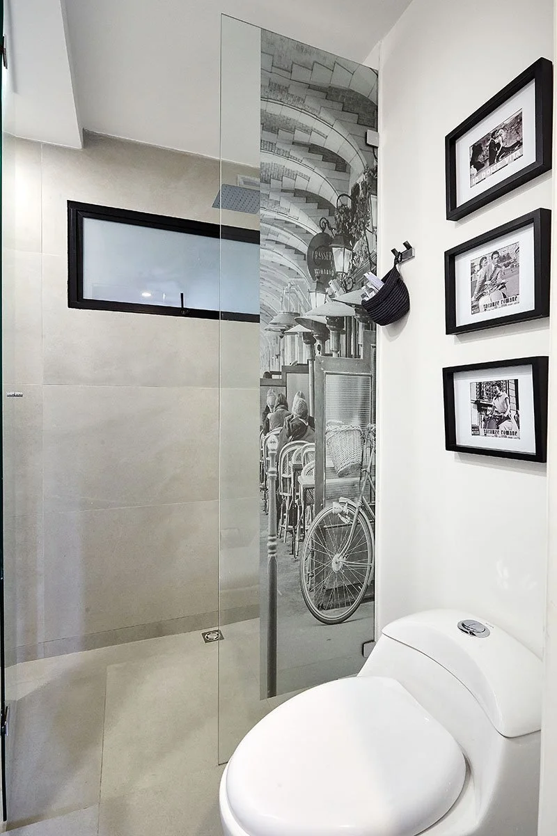 Baño con ducha de vidrio, un inodoro blanco, cuadros decorativos en la pared y una ventana pequeña.