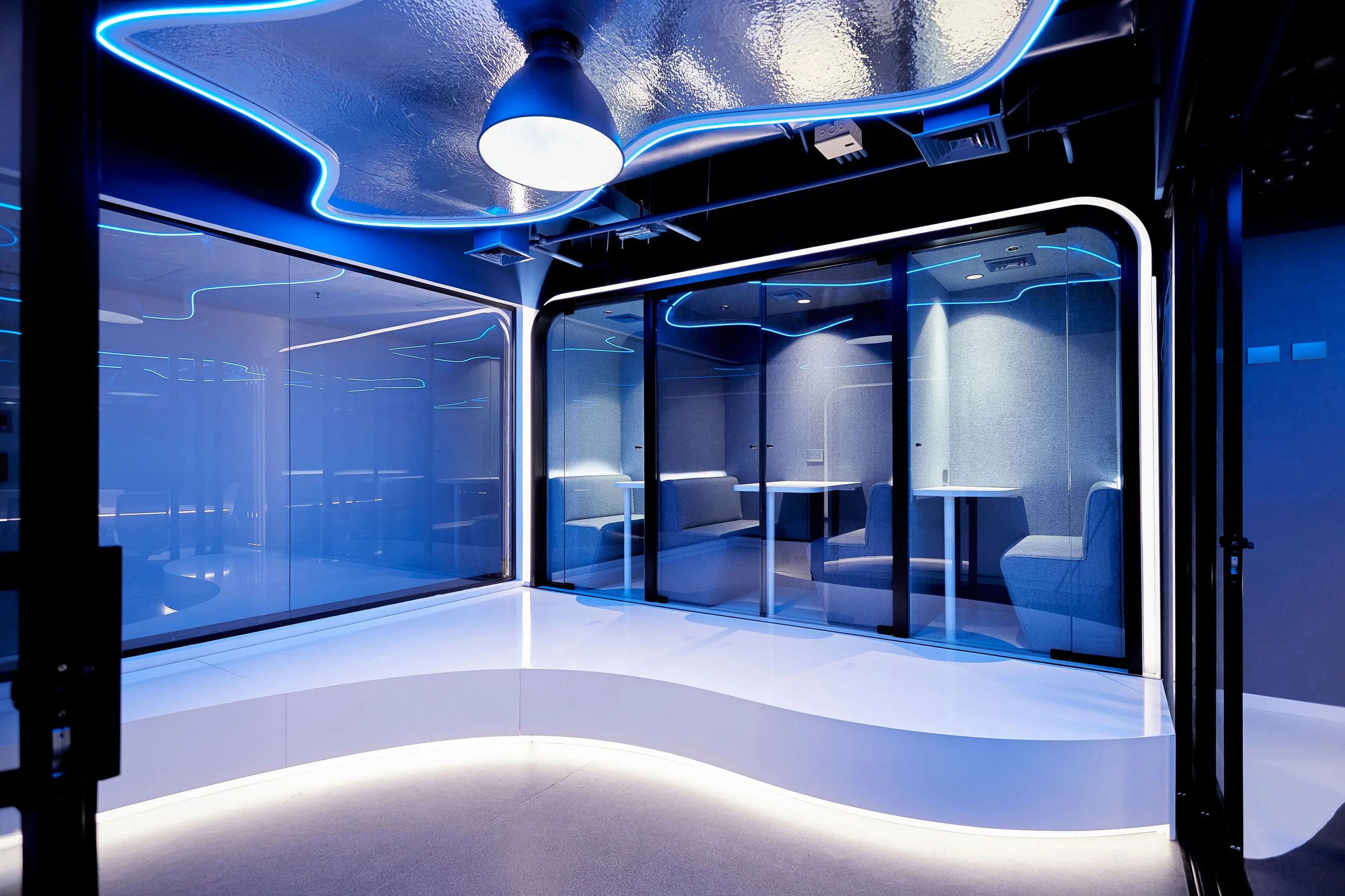Interior de una sala moderna con paredes de cristal y iluminación en tonos azules, con mesas y sillas blancas.