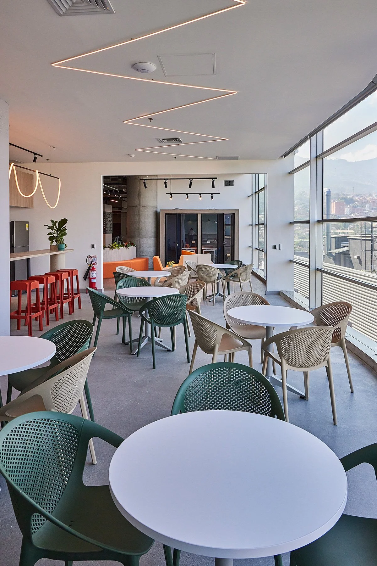 Interior de un espacio moderno con mesas redondas blancas y sillas verdes y beige, ventanas grandes con vista a la ciudad, decoración minimalista y iluminación moderna en el techo.
