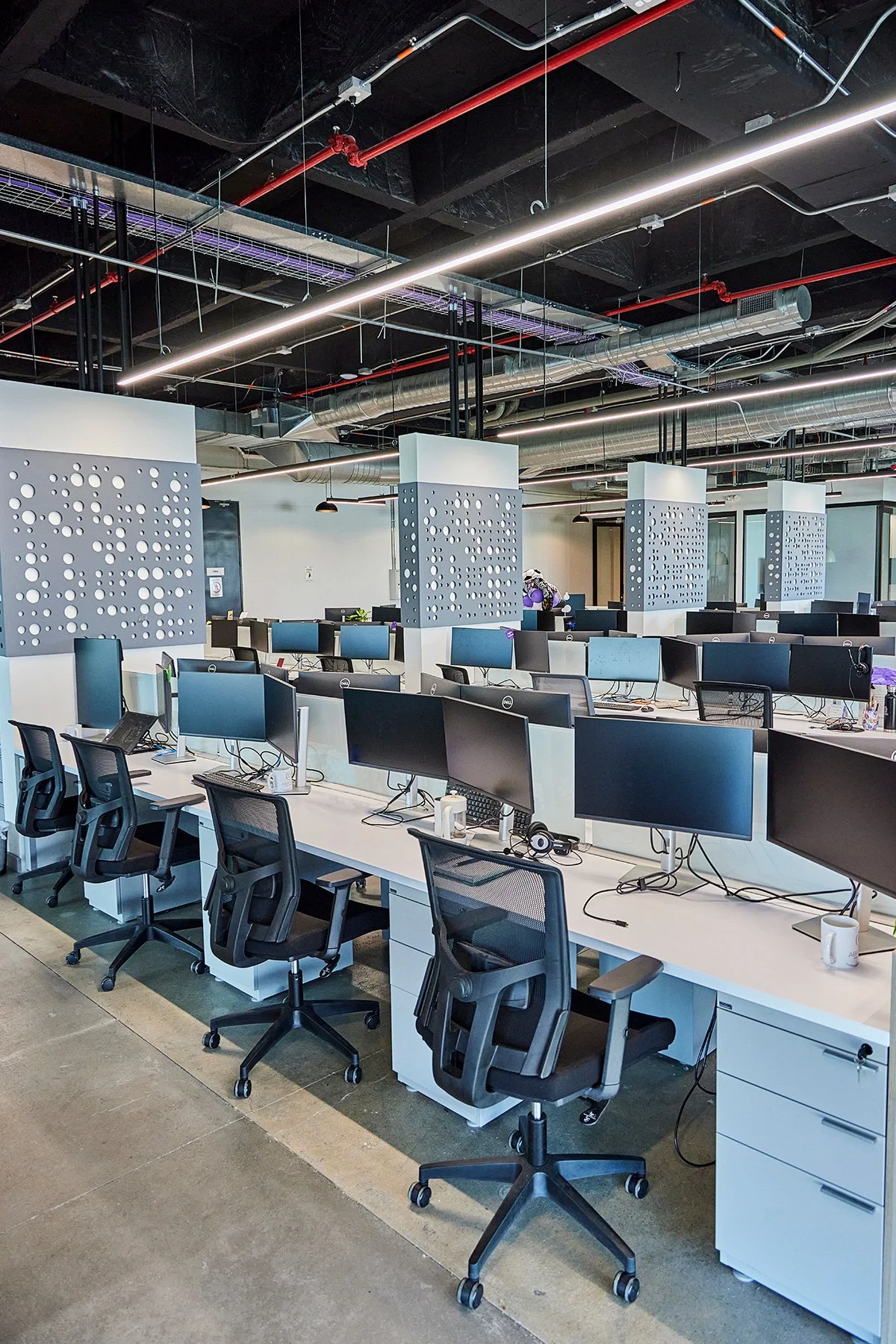 Oficina moderna con escritorios y monitores, sillas ergonómicas y divisores decorativos en las estaciones de trabajo, con un techo expuesto con ductos y tuberías de iluminación.