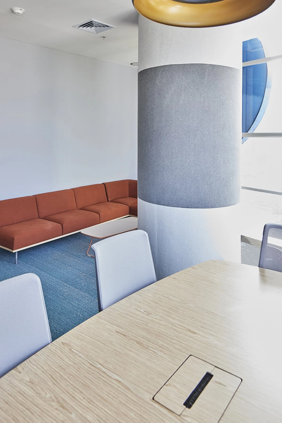 Área de vestíbulo o sala de espera con sofá naranja, mesa blanca, silla blanca, y una pared con columna cubo de tela gris, ventana con vista al exterior, y cielo despejado.