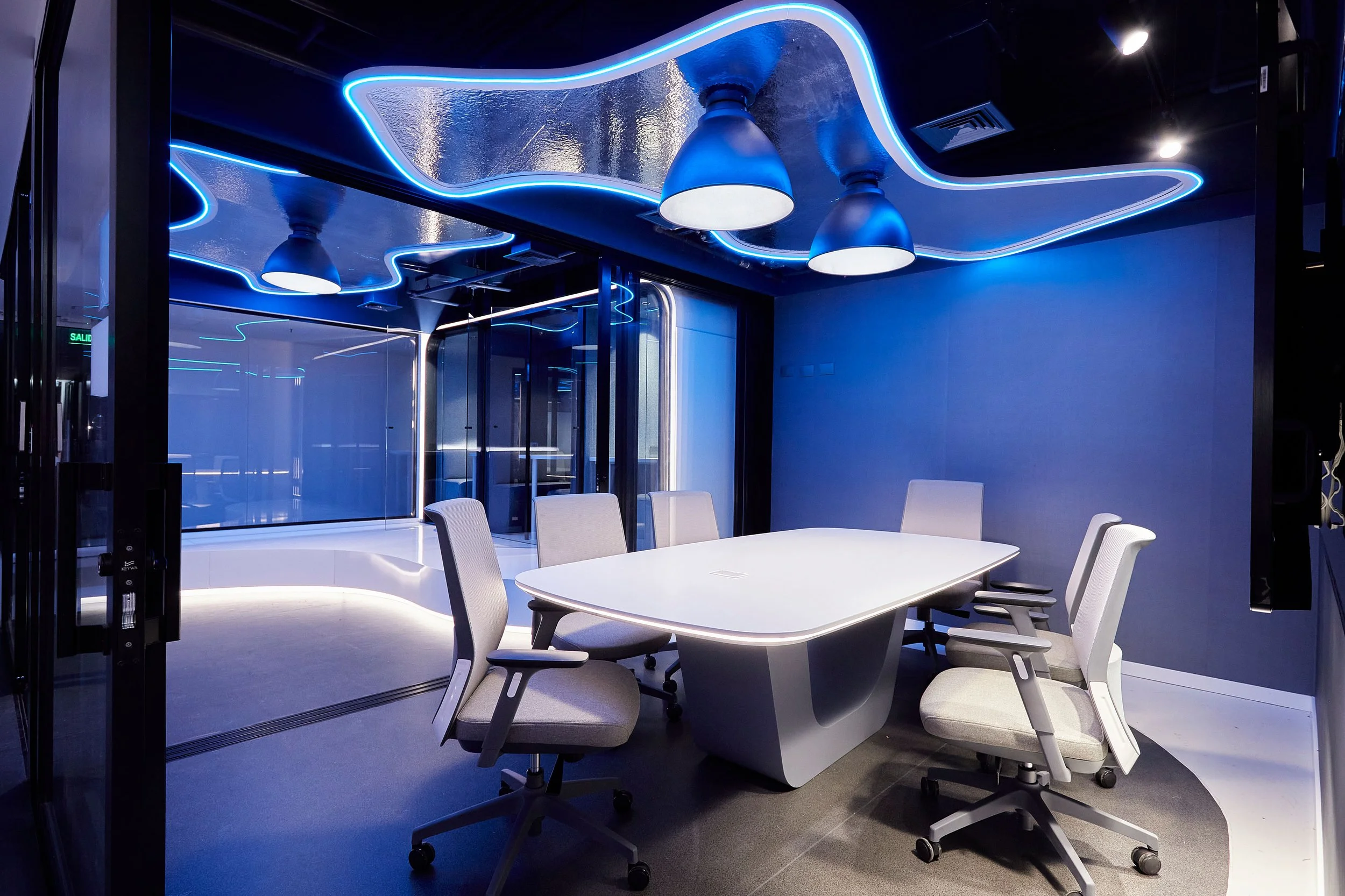 Sala de reuniones moderna con mesa blanca y sillas grises, iluminación de luces LED azules en el techo y paredes de vidrio reflectante.