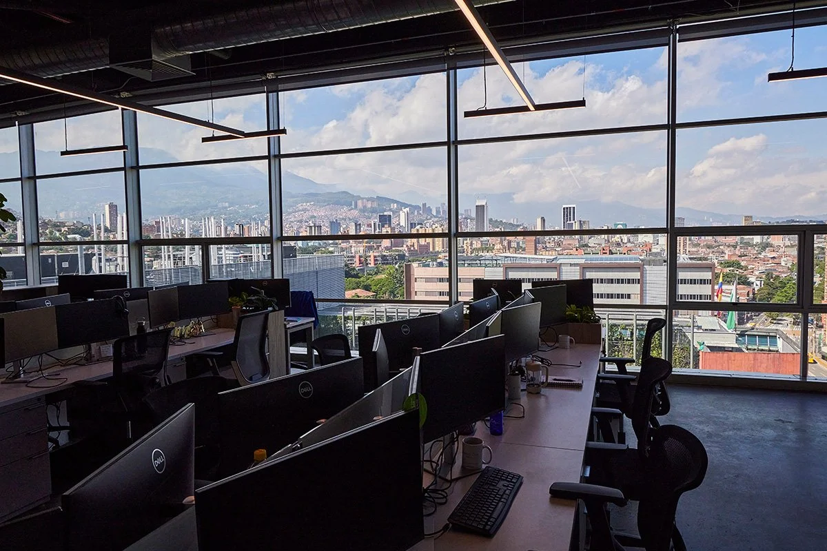 Oficina moderna con escritorios y monitores, vista panorámica de la ciudad y montañas al fondo.