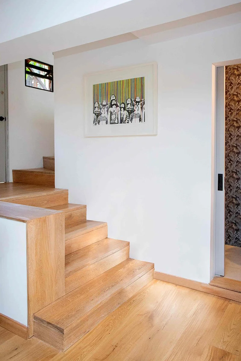 Escalera de madera en una casa moderna con paredes blancas, un cuadro en la pared y una ventana con vista al exterior.