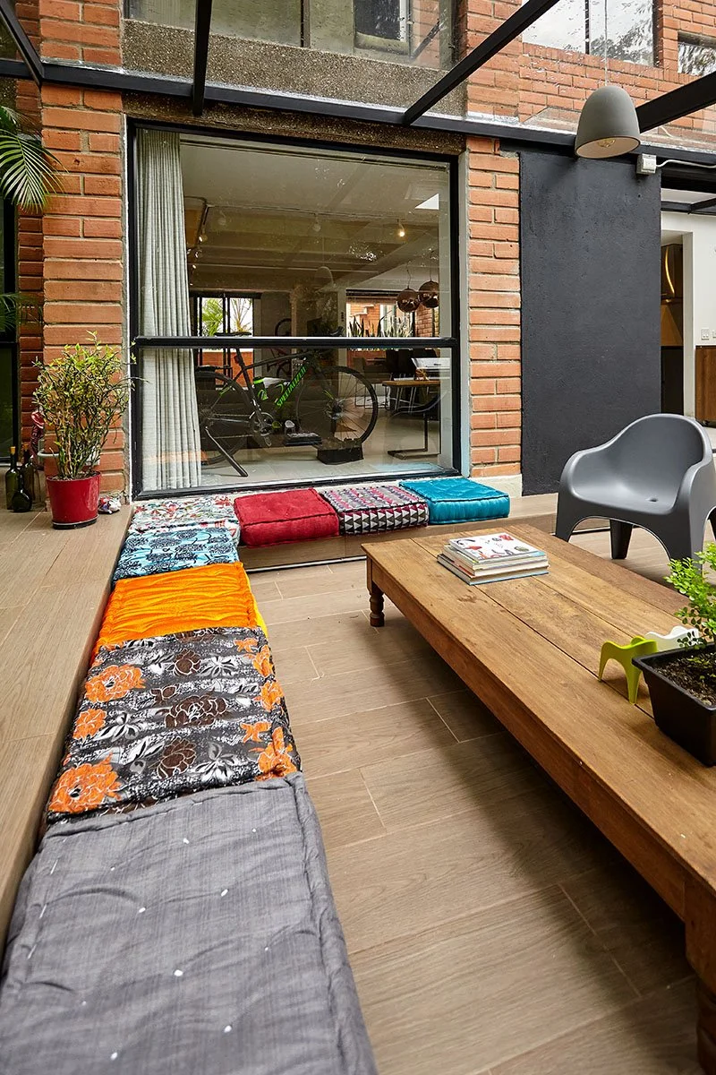 Interiores con sillón gris, mesa de madera, almohadones de colores y plantas decorativas, con vista a una habitación con bicicleta estática.