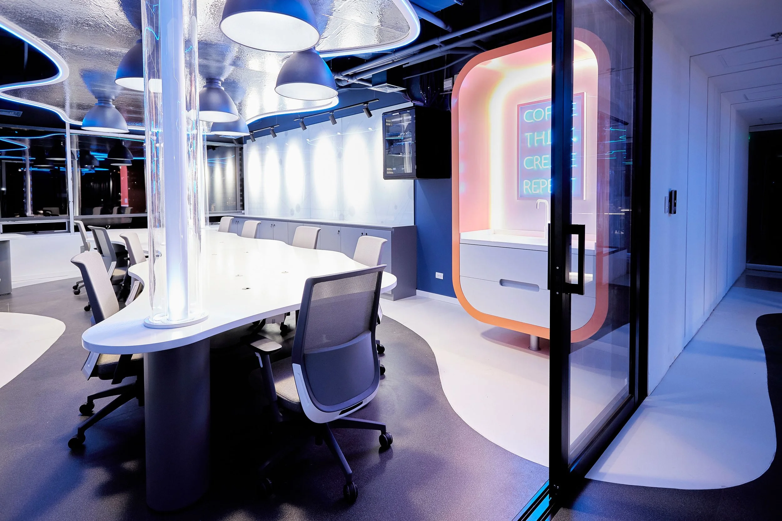 Sala de juntas moderna con mesa ovalada y sillas de oficina, iluminación de lámparas de color azul y paredes blancas, con una pared decorativa en tonos pastel y un cartel luminoso en el fondo que dice 'Coffee, Think, Create, Repeat'.