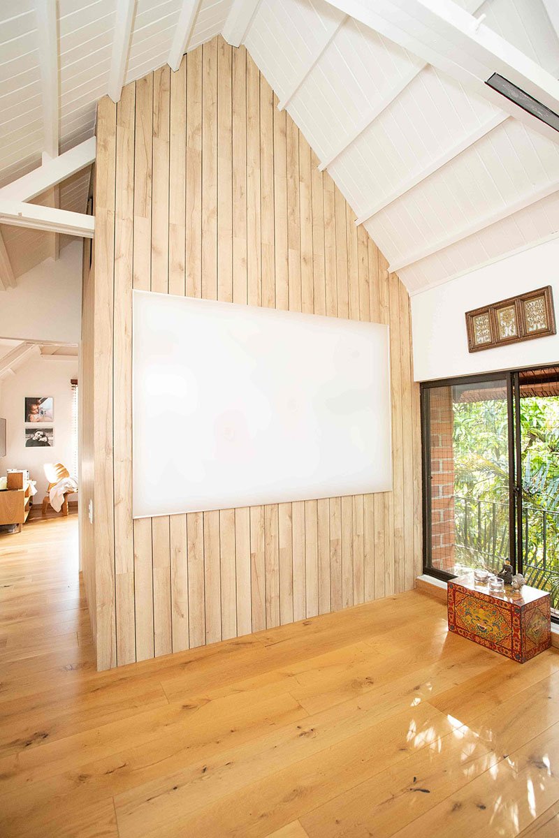 Interiores de una sala con paredes de madera clara, techo con vigas blancas, piso de madera y una ventana grande con vista a un balcón con plantas verdes.