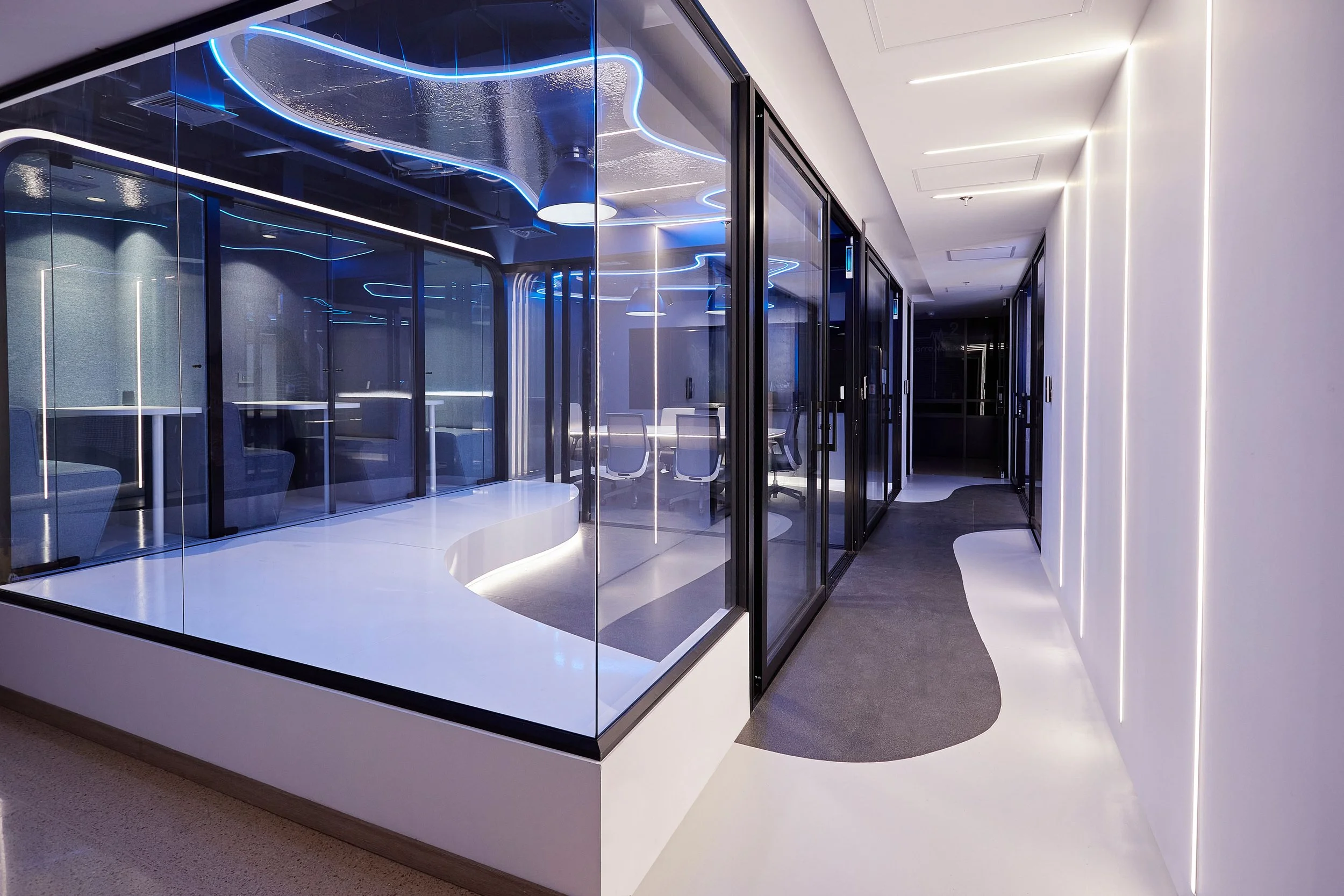 Pasillo moderno con paredes blancas, iluminación LED y una sala con paredes de vidrio y mobiliario en azul y gris.