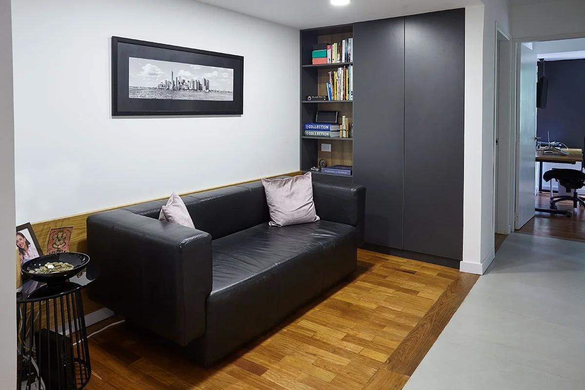 Sala de estar con sofá negro de cuero, estantería de colores oscuros llena de libros, cuadro en la pared y piso de madera.