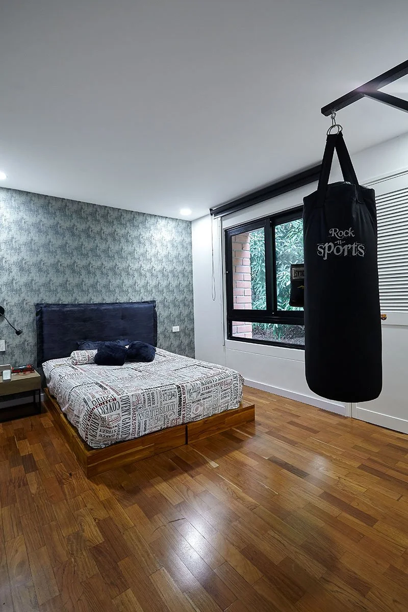 Habitación con cama, mesita de noche, pared decorativa, ventana con vista a áreas verdes y saco de boxeo colgado del techo.