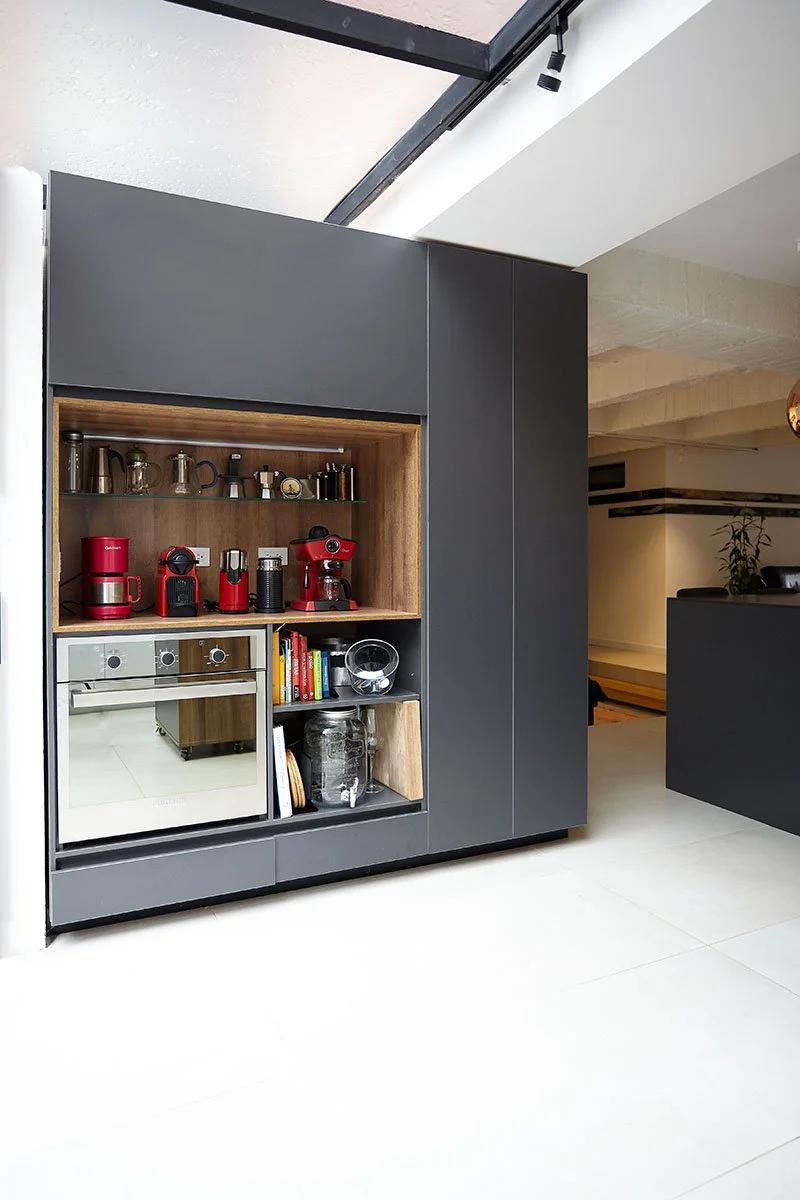 Gabinete negro con electrodomésticos y utensilios de cocina, incluyendo una máquina de café, una máquina de espresso, libros de cocina, y algunos vasos y tazas, en una cocina moderna.