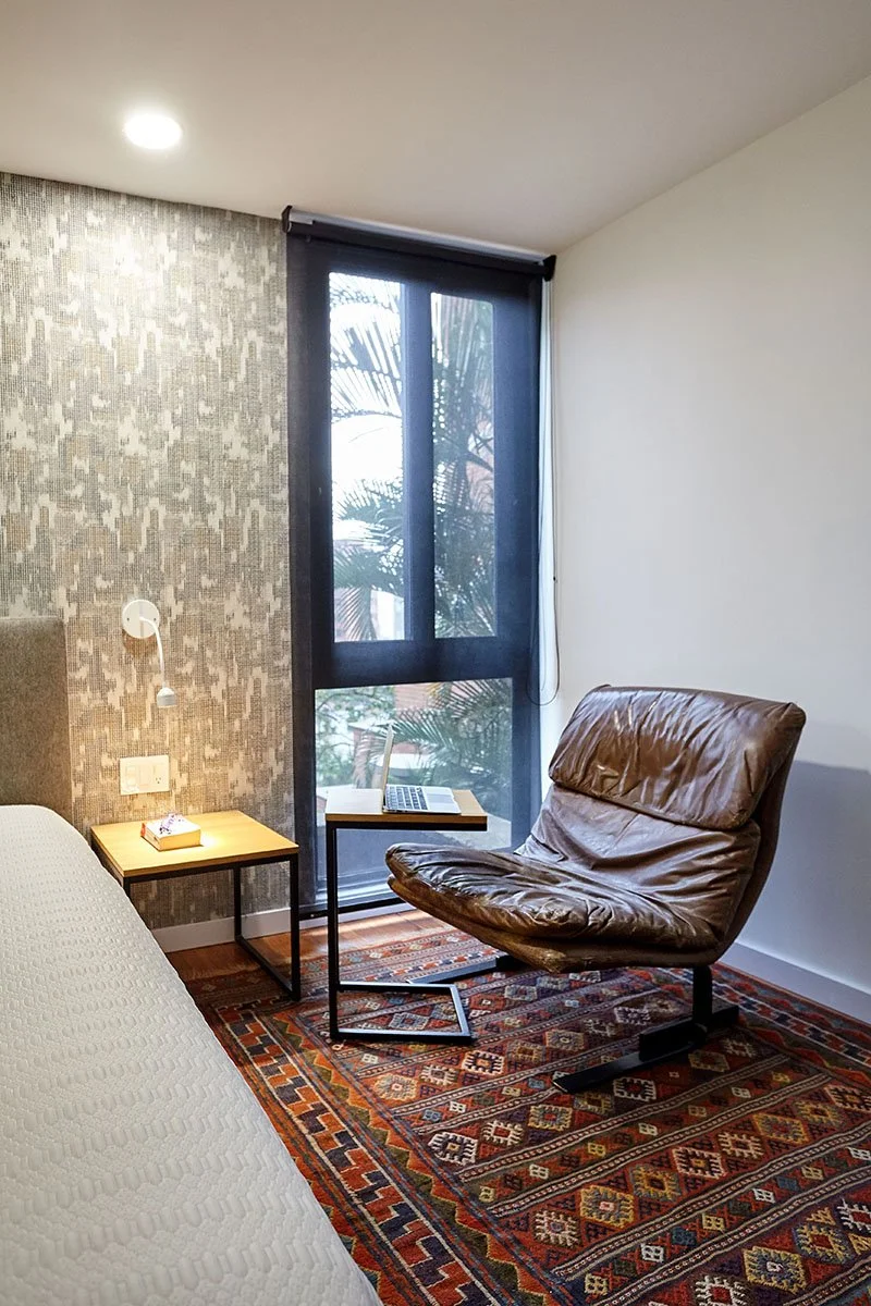 Habitación con una cama, una mesa con una laptop, una silla de cuero marrón y una ventana con cortina negra.