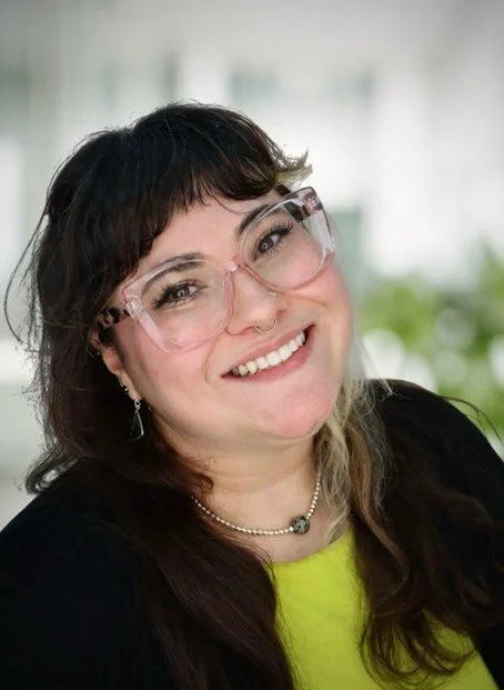 A smiling woman with glasses, dark hair, and a septum piercing, wearing a black top and yellow shirt indoors.
