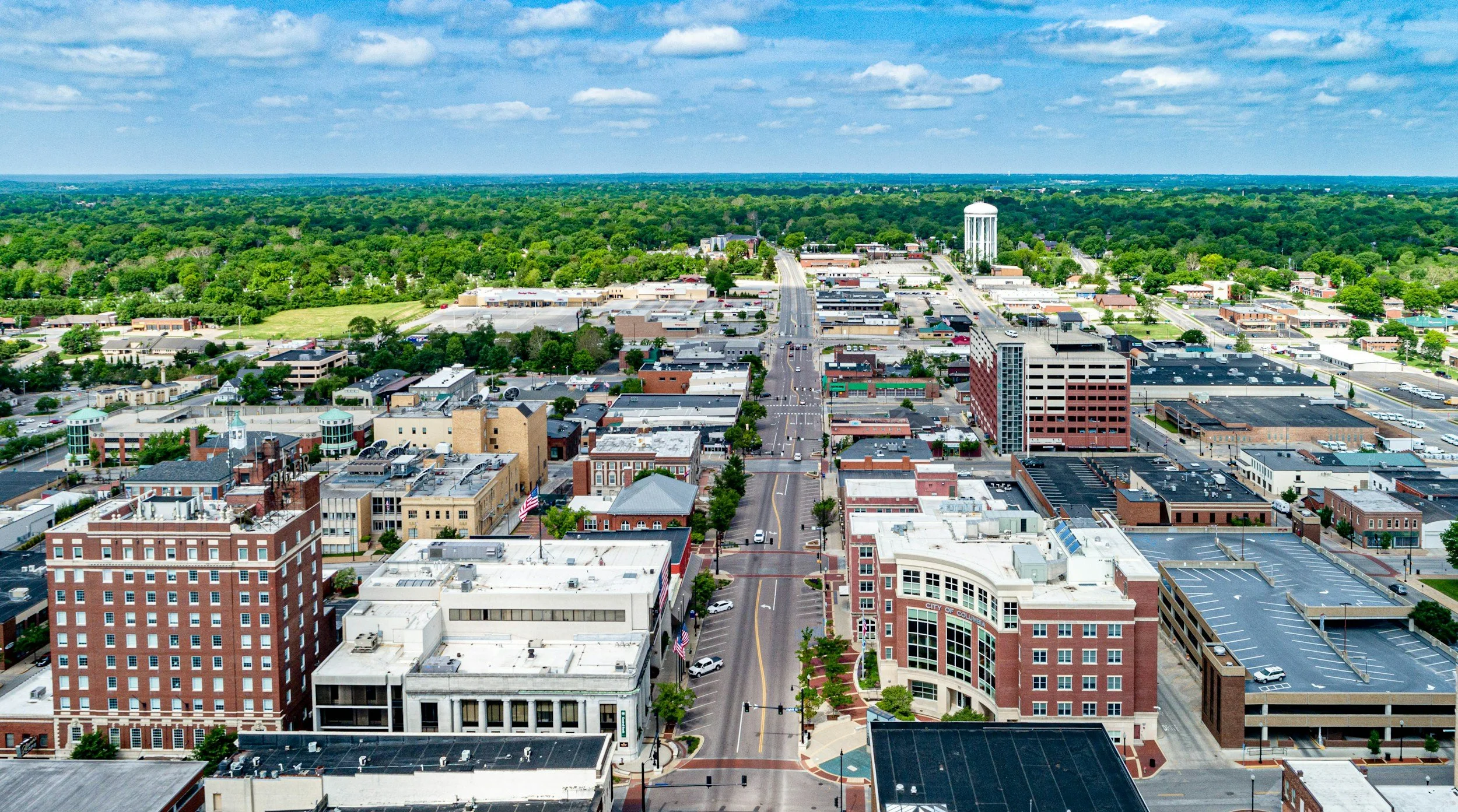 Aerial view of a city with streets, buildings, and a water tower in the background, surrounded by green trees and open land under a partly cloudy sky.