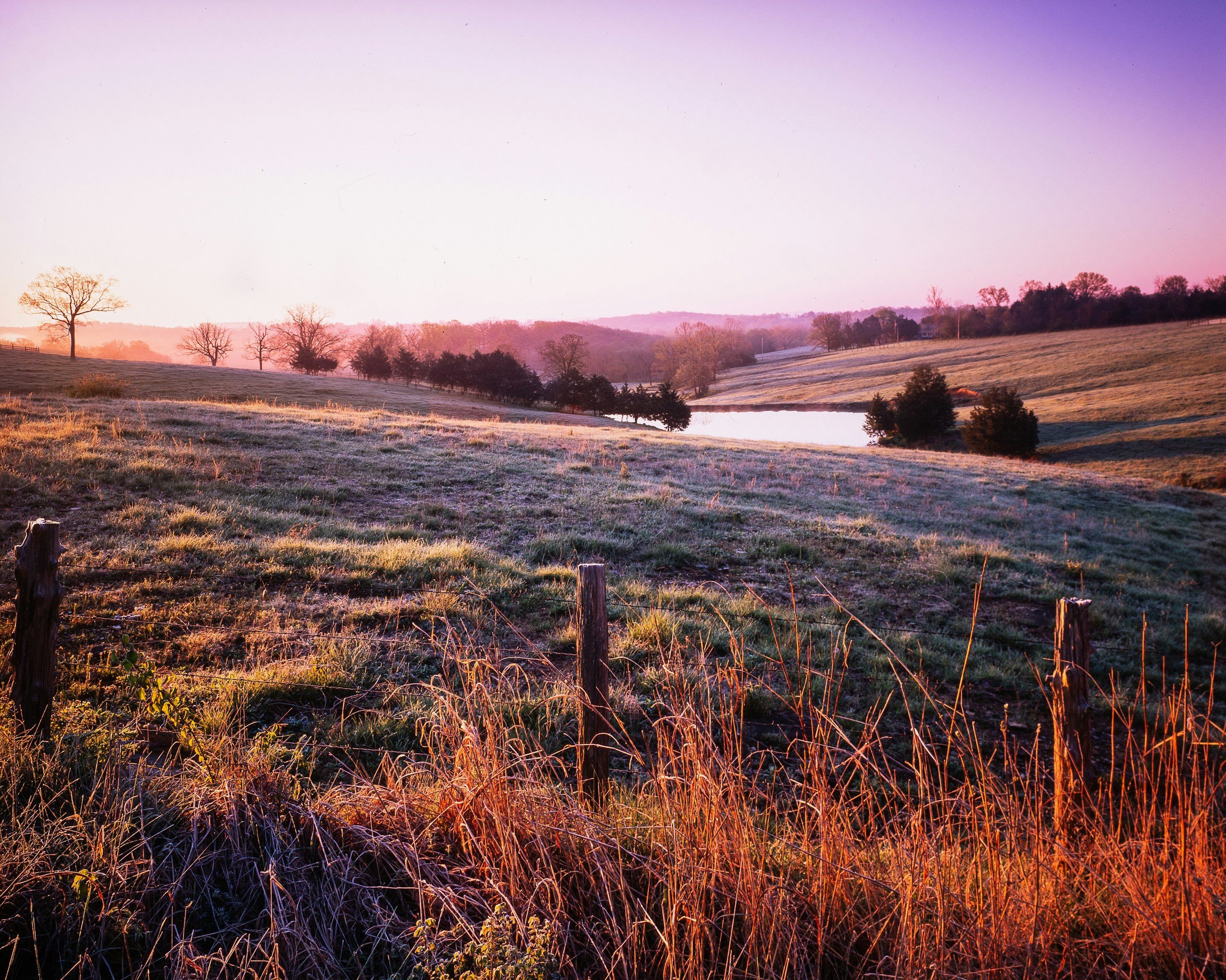 Sunrise over rolling hills with trees, a fence in the foreground, and a small body of water in the distance.
