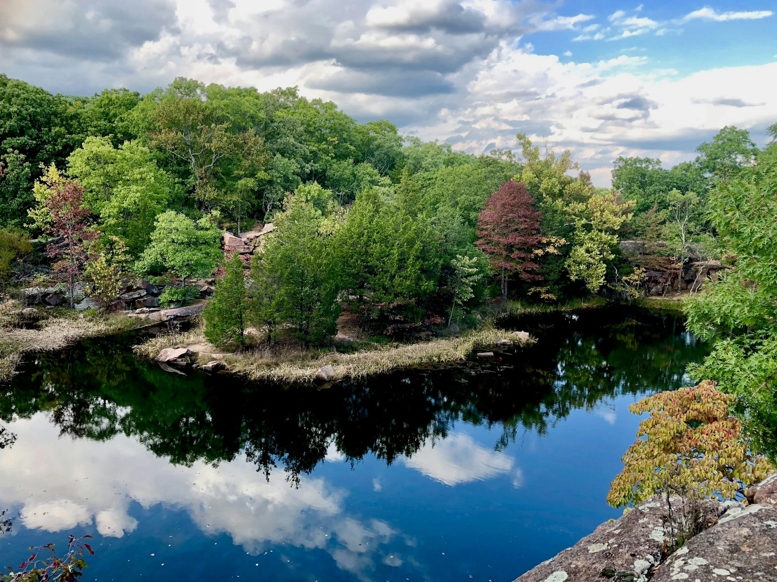 A peaceful landscape with a calm river reflecting the green trees and partly cloudy sky. Rocks and foliage border the river, and a hill covered with dense trees rises in the background.
