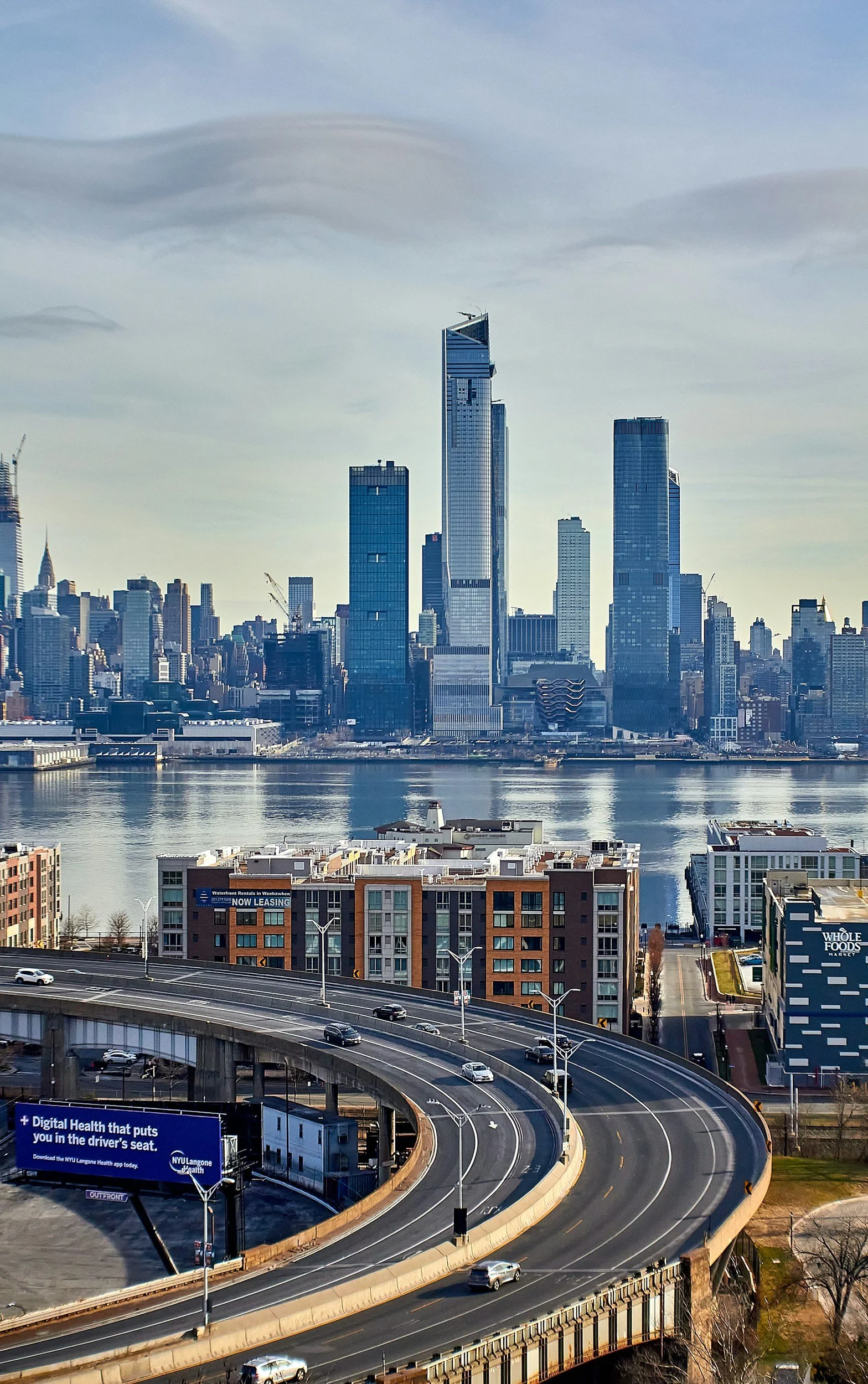 Downtown Manhattan skyline across the river with tall glass skyscrapers, including the Central Park Tower, and a highway with moving cars in the foreground.