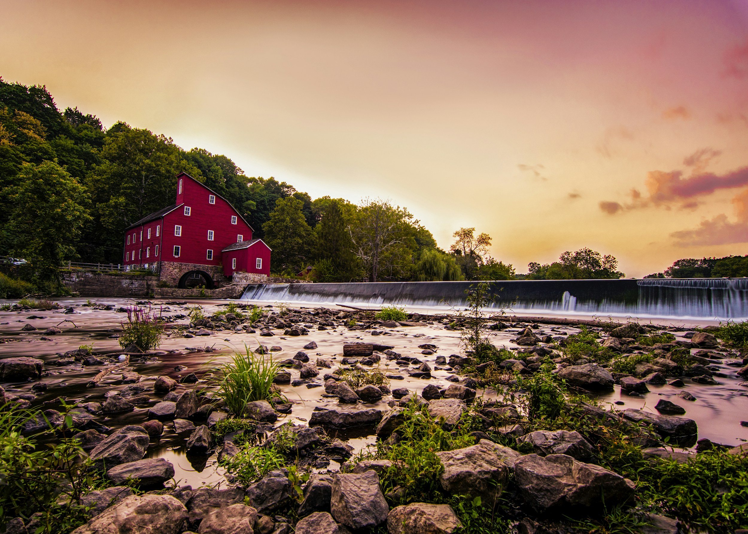 A red historic mill building beside a flowing river at sunset, surrounded by trees and rocks in the foreground.