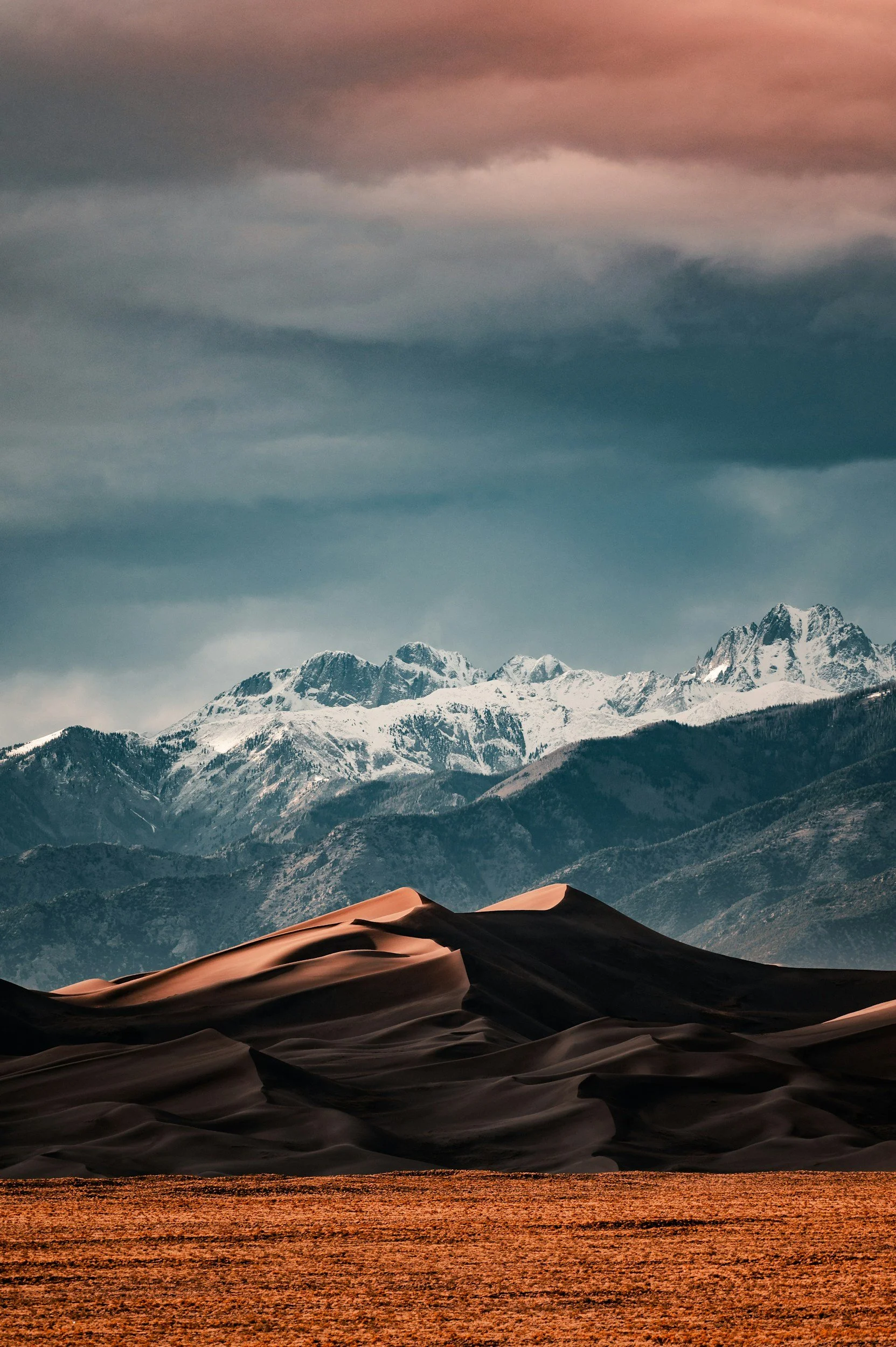 Scenic landscape featuring sand dunes in the foreground, mountains with snow-capped peaks in the background, and a cloudy sky overhead.