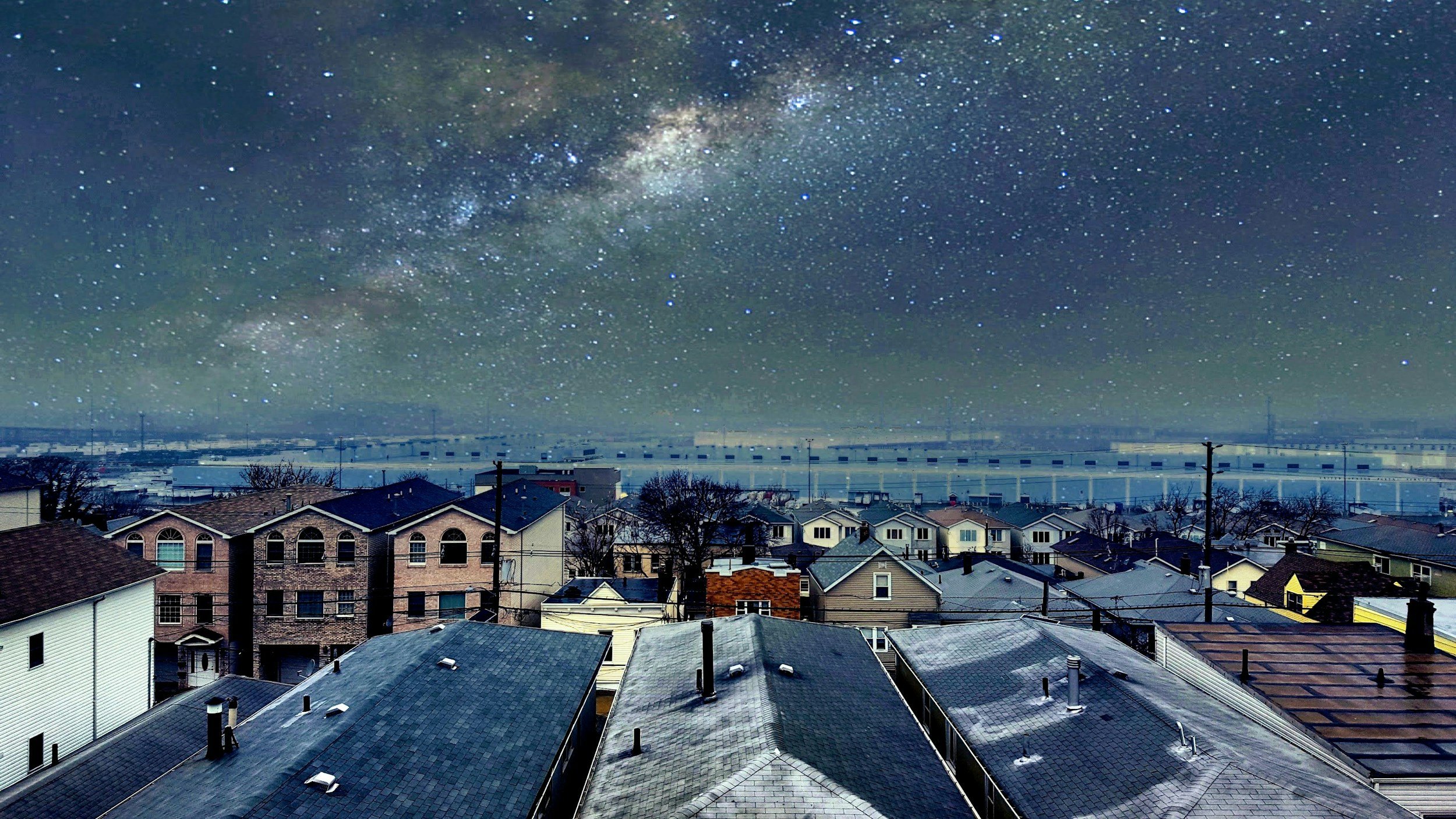 Night view of a suburban neighborhood with rooftops, under a starry sky showcasing the Milky Way galaxy.