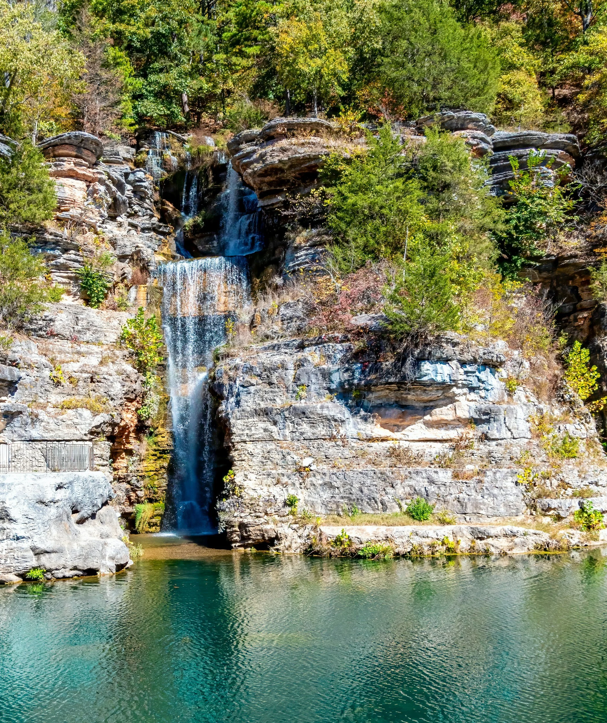A scenic landscape featuring a multi-tiered waterfall cascading down rocky cliffs into a calm river, with lush green trees and foliage in the background.