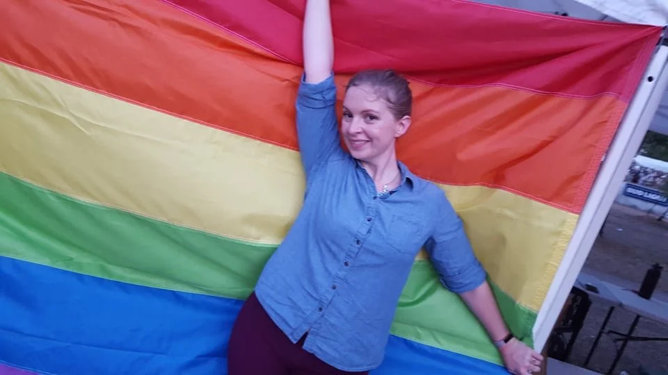 A woman with a ponytail smiling and posing with one arm raised, standing in front of a rainbow pride flag.