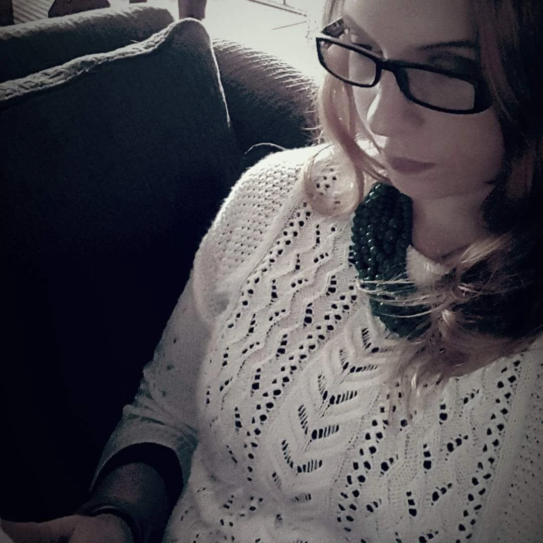 A woman with shoulder-length hair, wearing glasses, a white lace sweater, and a beaded necklace, sitting on a dark couch in a room with natural light coming through a window.