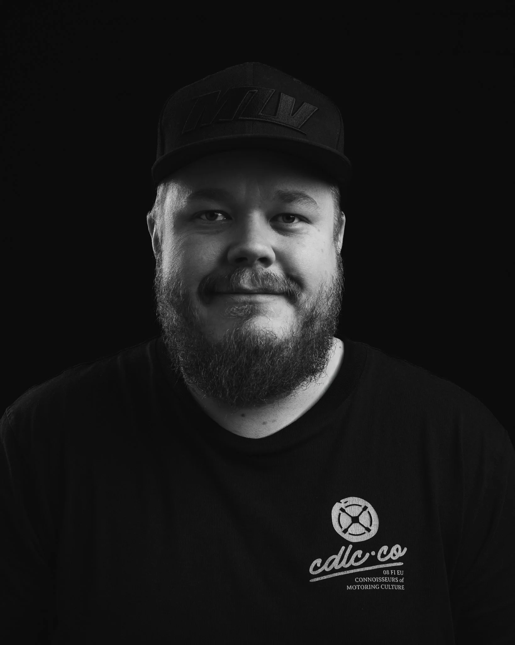 Hieno musta baseball lippis ja musta T-paita, joissa molemmissa on valkoinen logo ja teksti, ja henkilön kasvot näkyvät osittain.