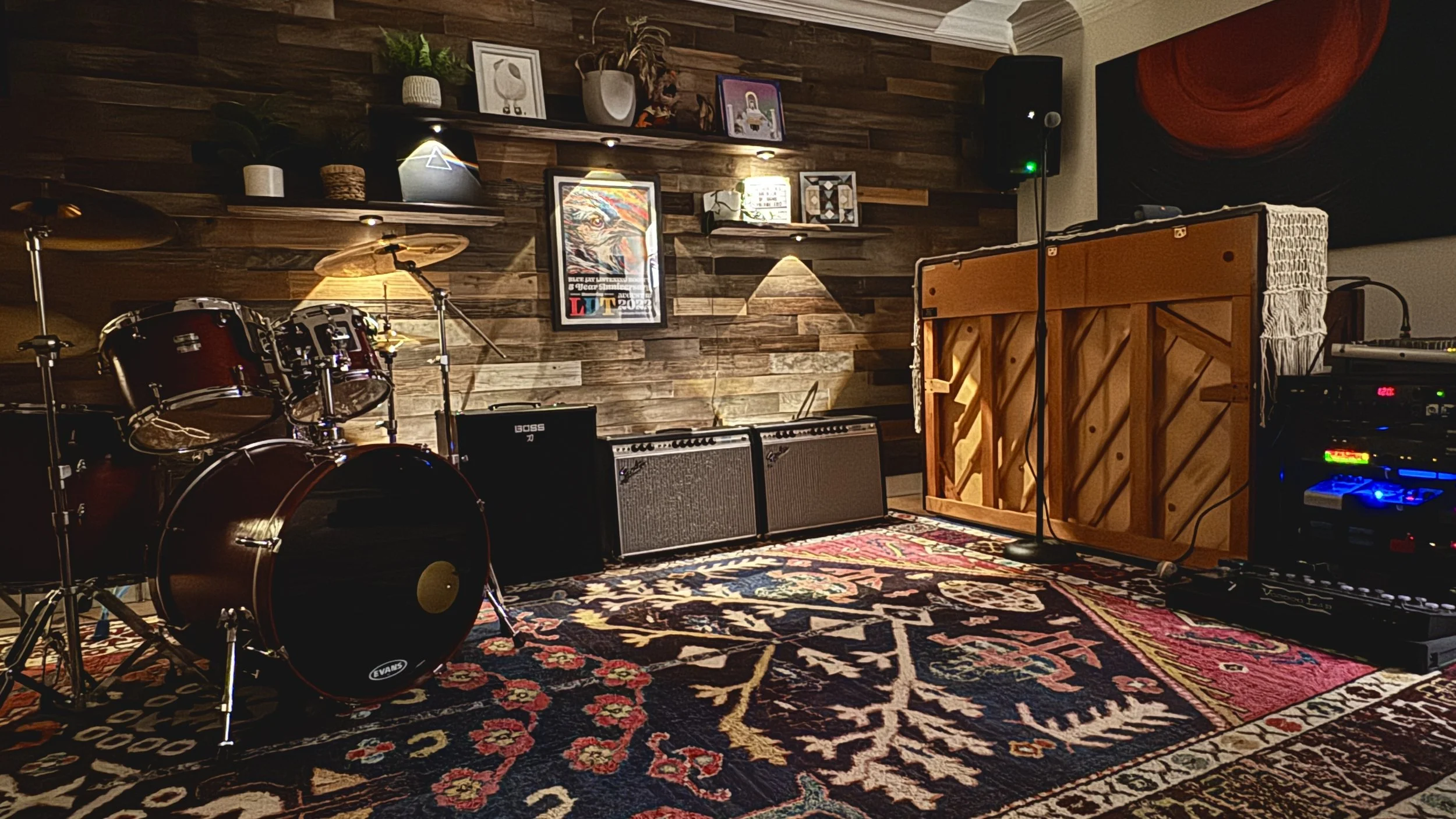 A music rehearsal space with drums, guitar amplifiers, and audio equipment, set against a wooden wall with framed artwork and potted plants, illuminated by warm lighting.