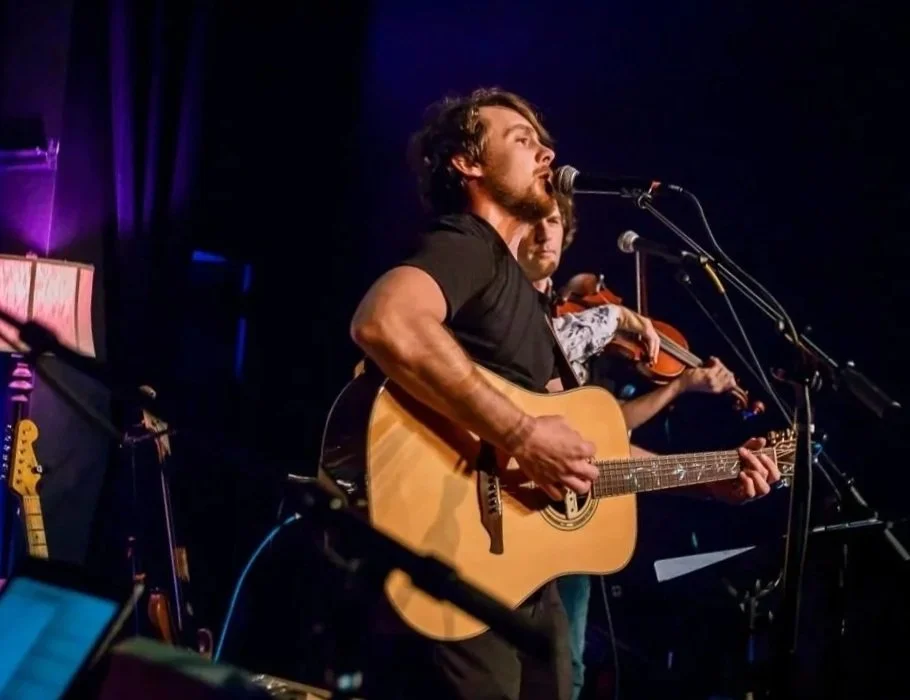 A musician with curly hair and a beard playing an acoustic guitar on stage, with a violinist in the background.