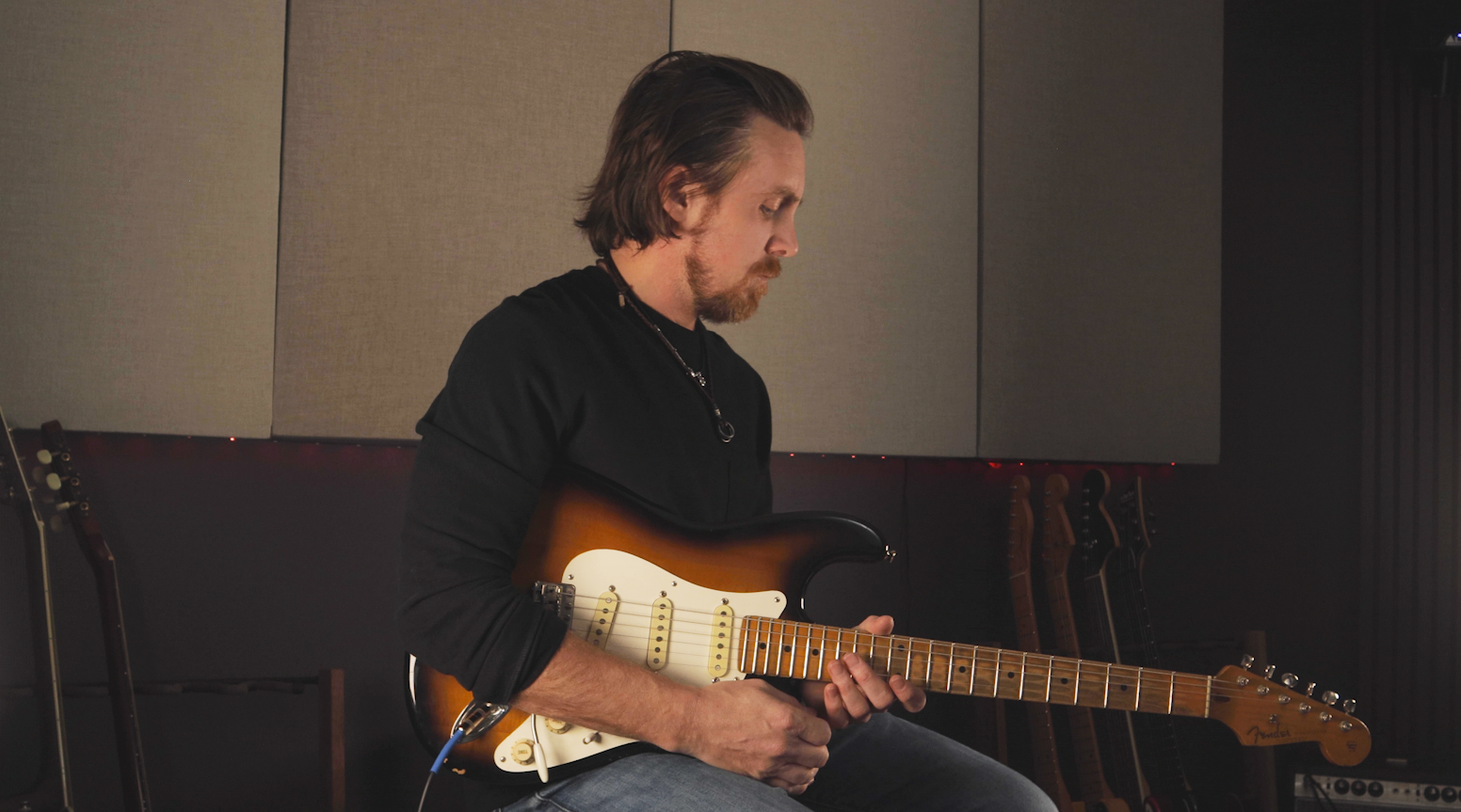 A man with shoulder-length brown hair and a beard playing an electric guitar in a music studio.