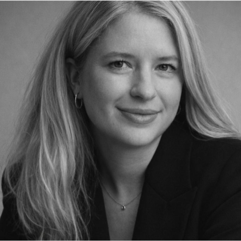 A black and white portrait of a woman with long hair, wearing earrings and a necklace, smiling slightly, dressed in a dark blazer.