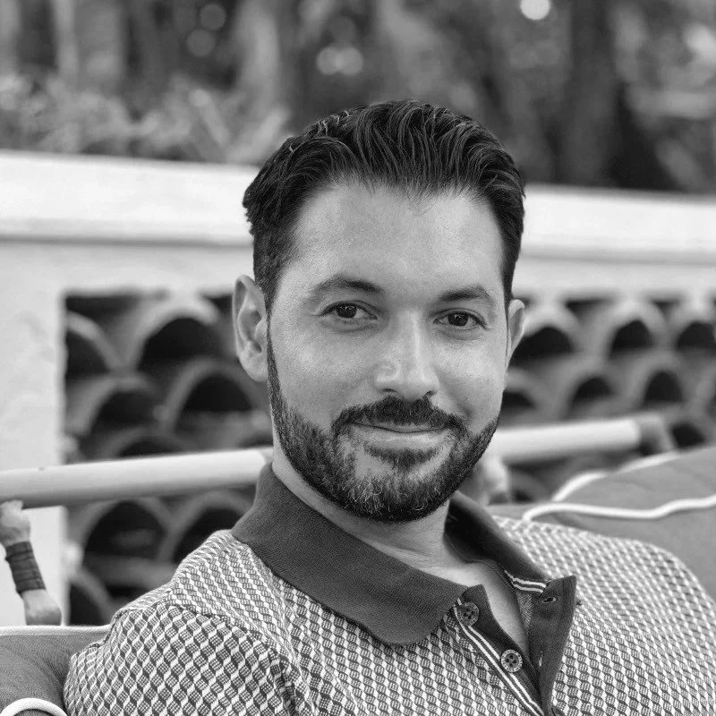 Black and white photo of a man with dark hair and a beard, smiling, sitting outdoors with a blurred background.