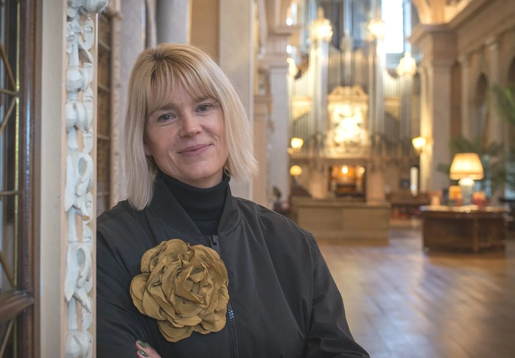 A woman with blonde hair wearing a black jacket and a large, gold-colored flower on her chest, standing in an elegant, ornate interior with chandeliers, wooden flooring, and decorative elements.