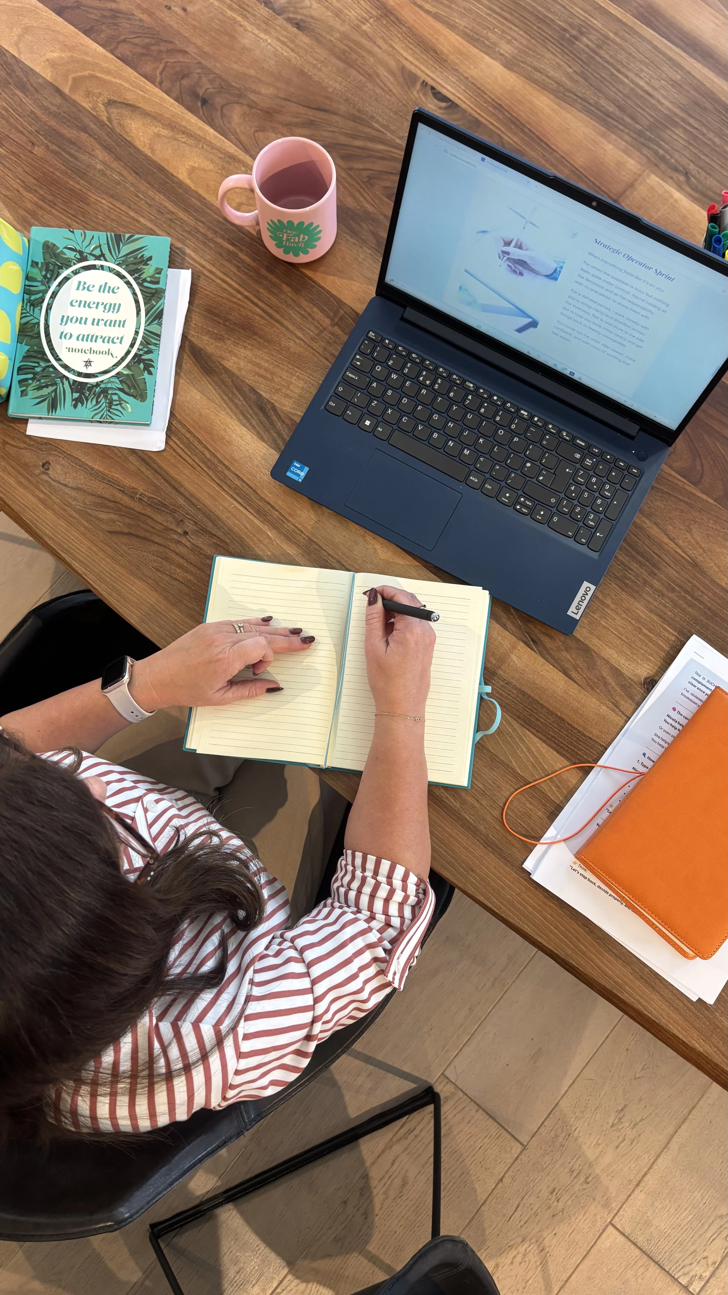 A woman sitting at a wooden table with a laptop, an open notebook, and various notebooks and papers. She is writing in the notebook with a pen. There is a pink mug with tea or coffee, a teal notebook with a motivational quote, and white and orange notebooks on the table.