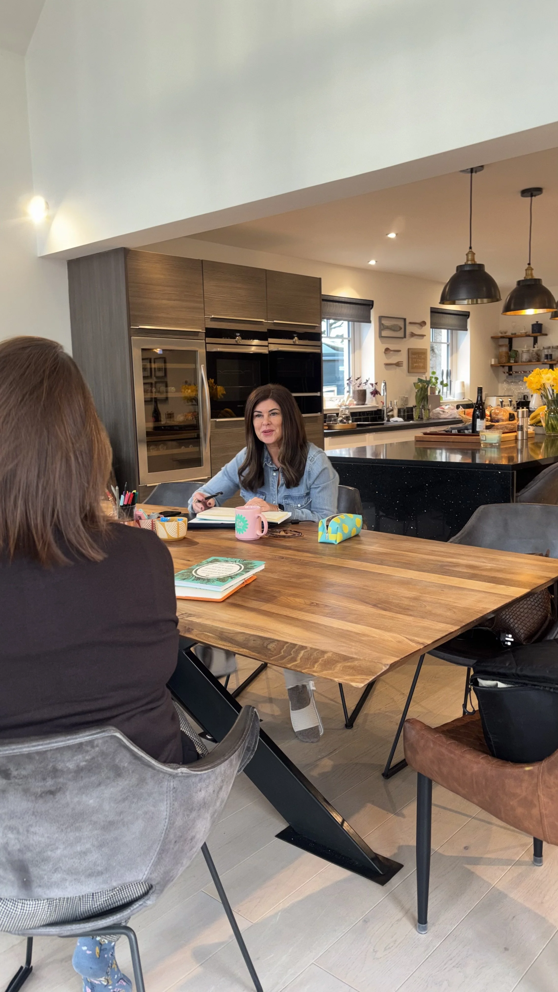 Two women are seated at a wooden table, engaged in conversation in a modern kitchen with kitchen appliances, windows, and decorative wall art in the background.