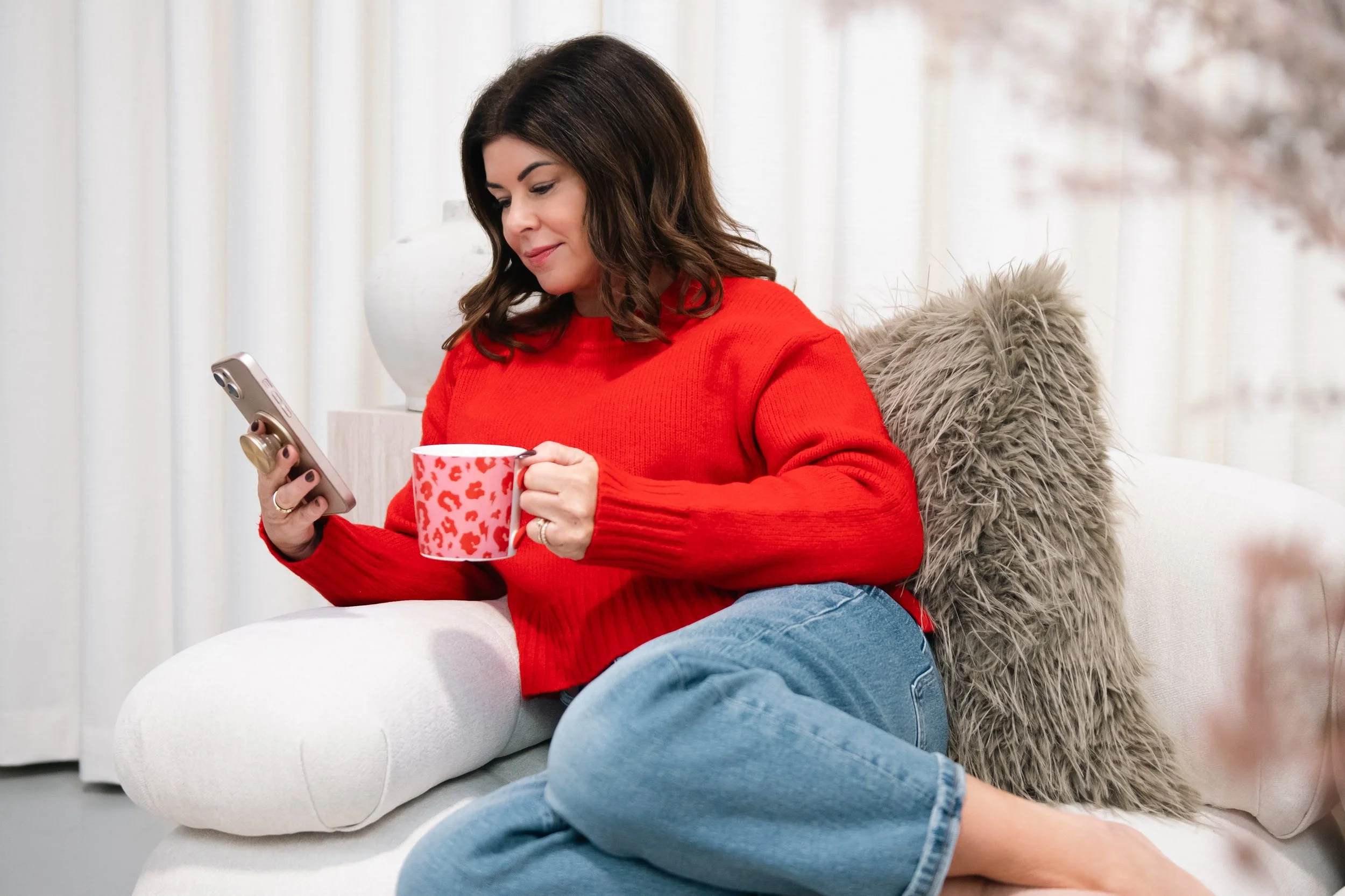 Woman sitting on a white sofa, holding a smartphone in one hand and a red patterned mug in the other, wearing a red sweater and blue jeans, with a fluffy pillow behind her, in a well-lit room with white curtains.