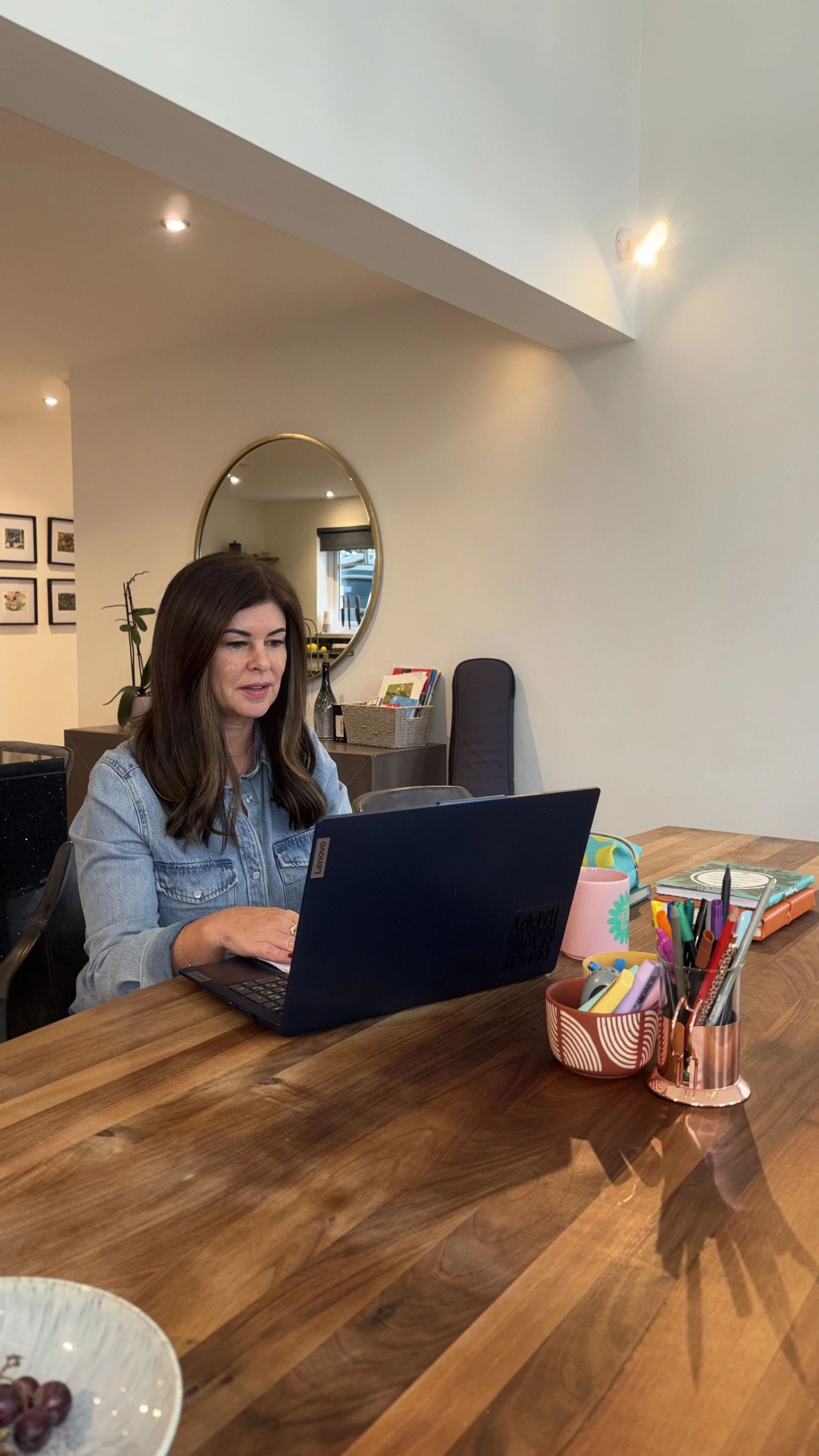 Woman with brown hair working on a laptop at a wooden table in a well-lit room with wall art, a mirror, and a plant in the background.