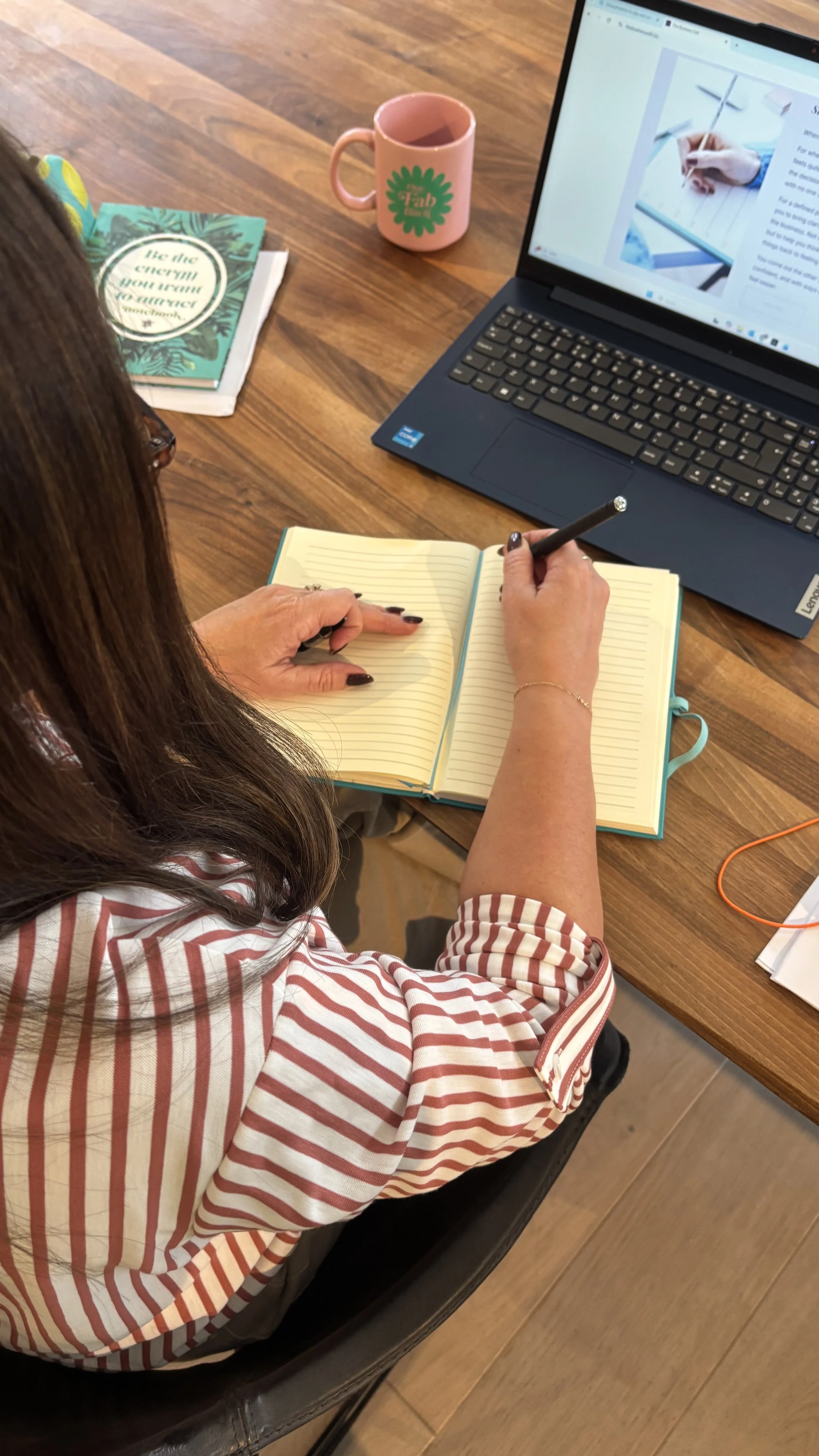 A woman with brown hair takes notes in a yellow notebook at a wooden table. There is a laptop, a pink mug, and a book on the table.