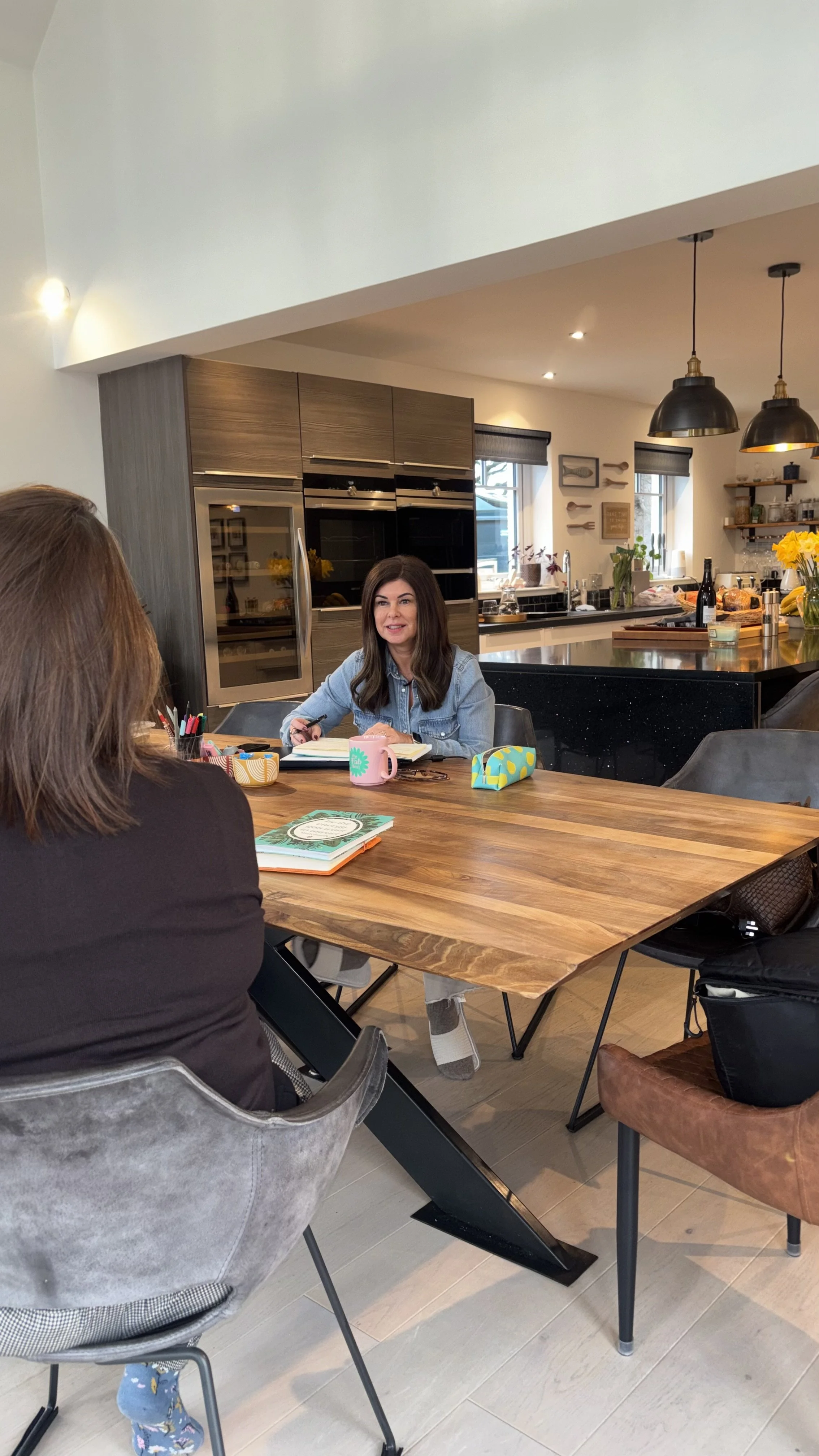 Two women having a discussion at a wooden dining table in a modern kitchen, with papers, notebooks, and colorful pens on the table.