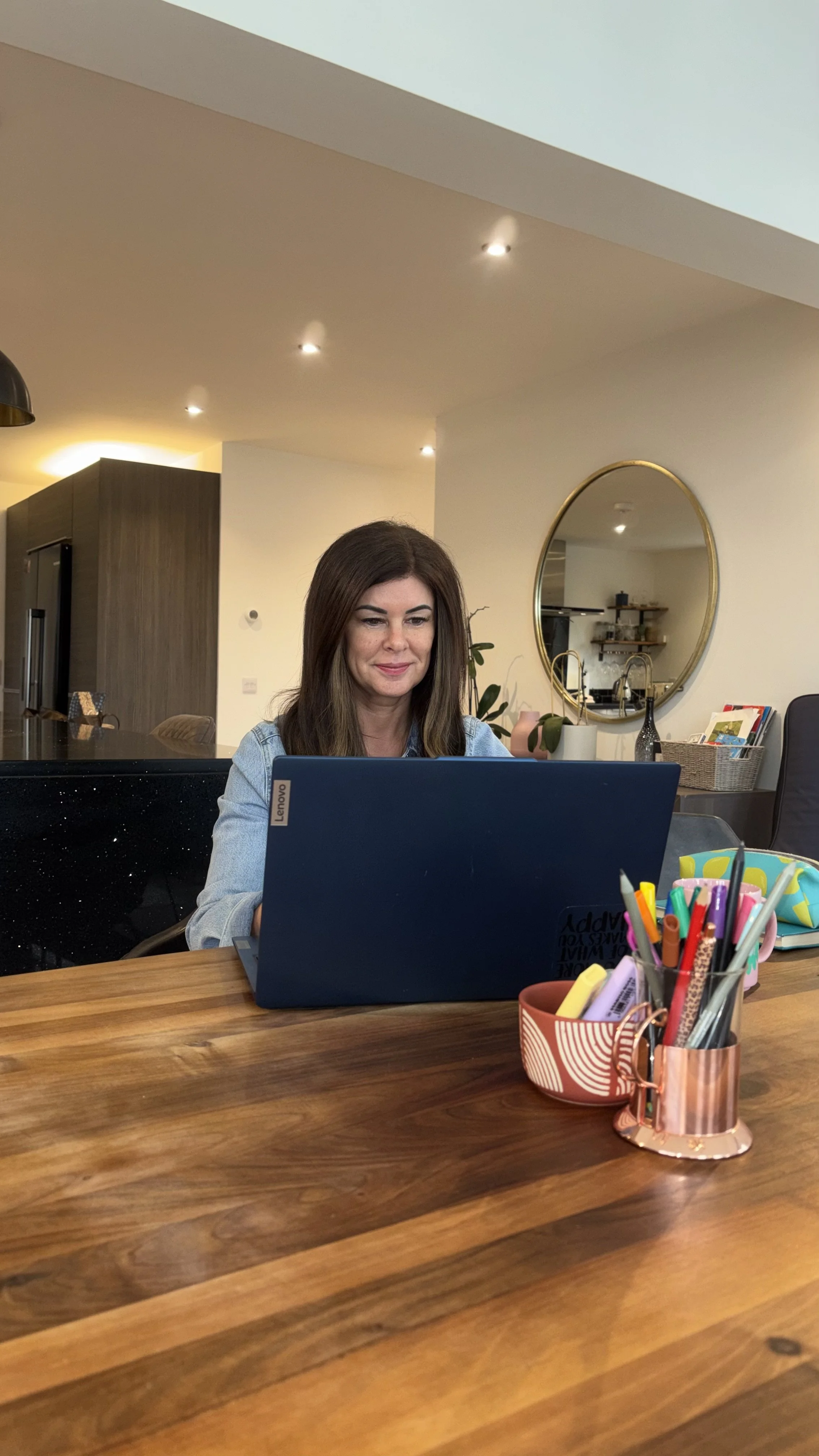 A woman with brown hair working on a Lenovo laptop at a wooden table in a modern home interior.