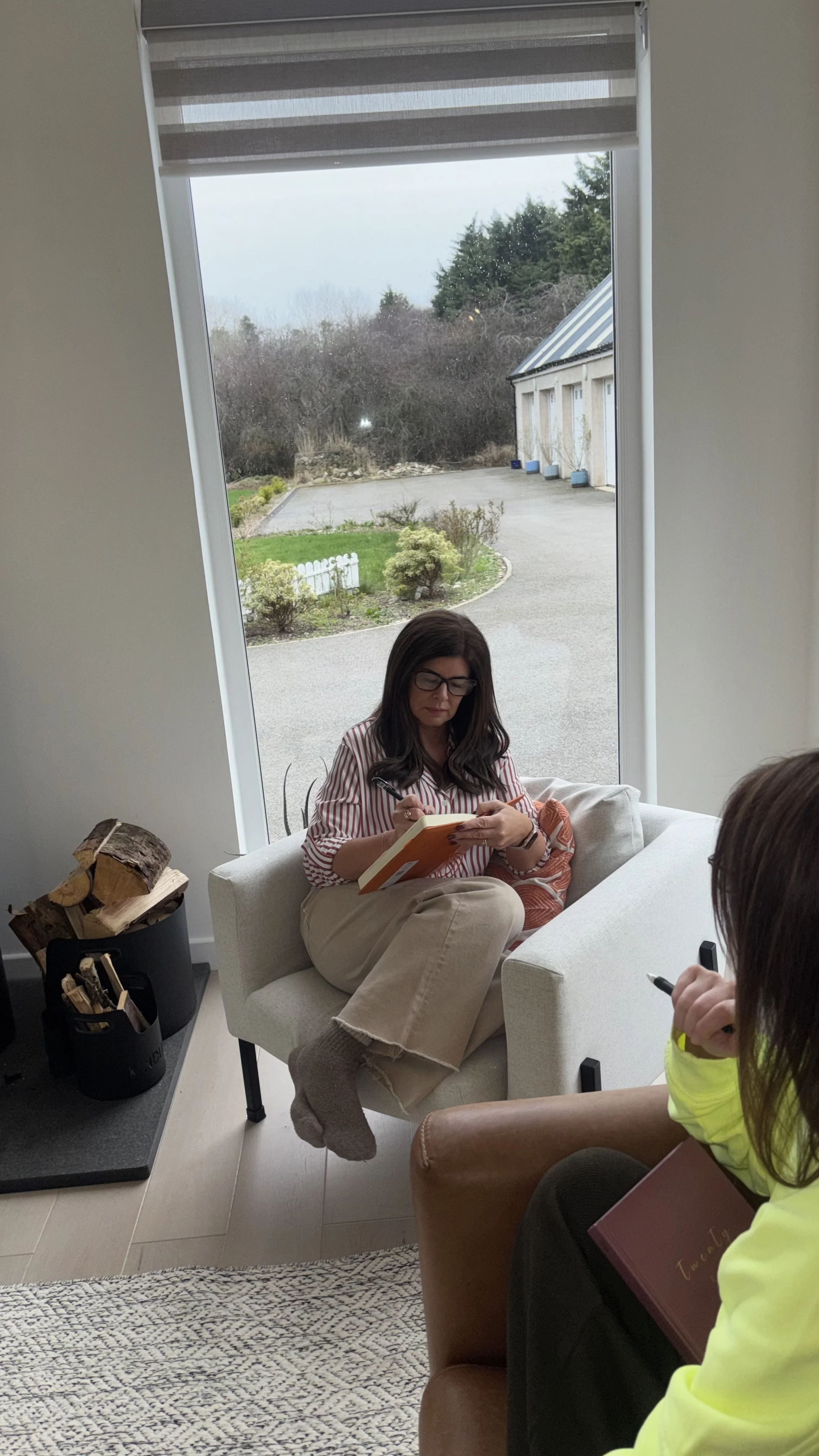 Woman sitting in a white armchair by a large window, reading a book or notebook, with a person in a yellow shirt sitting on a chair nearby.