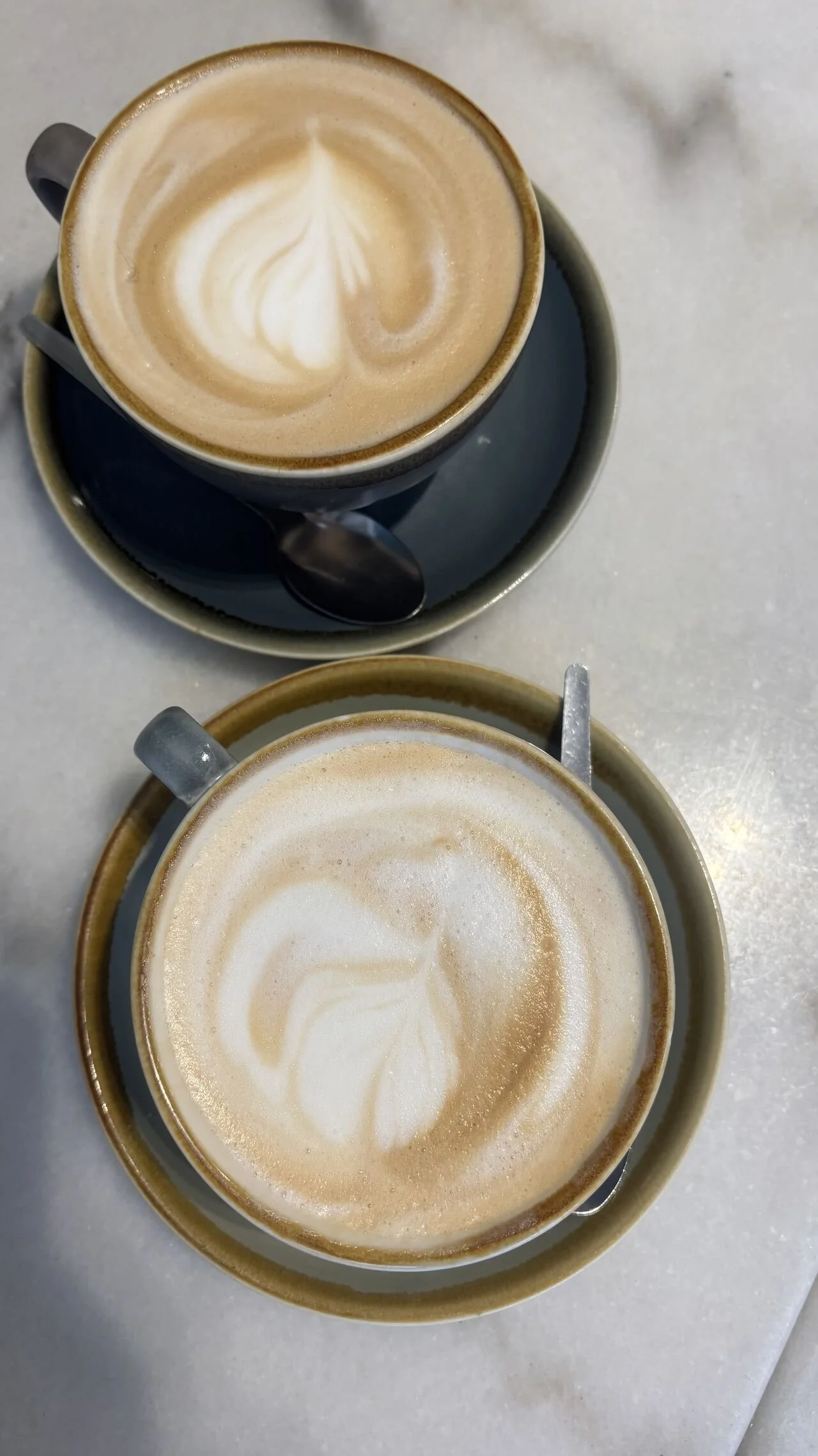 Two cups of coffee with latte art, served on saucers with small spoons, placed on a white surface.