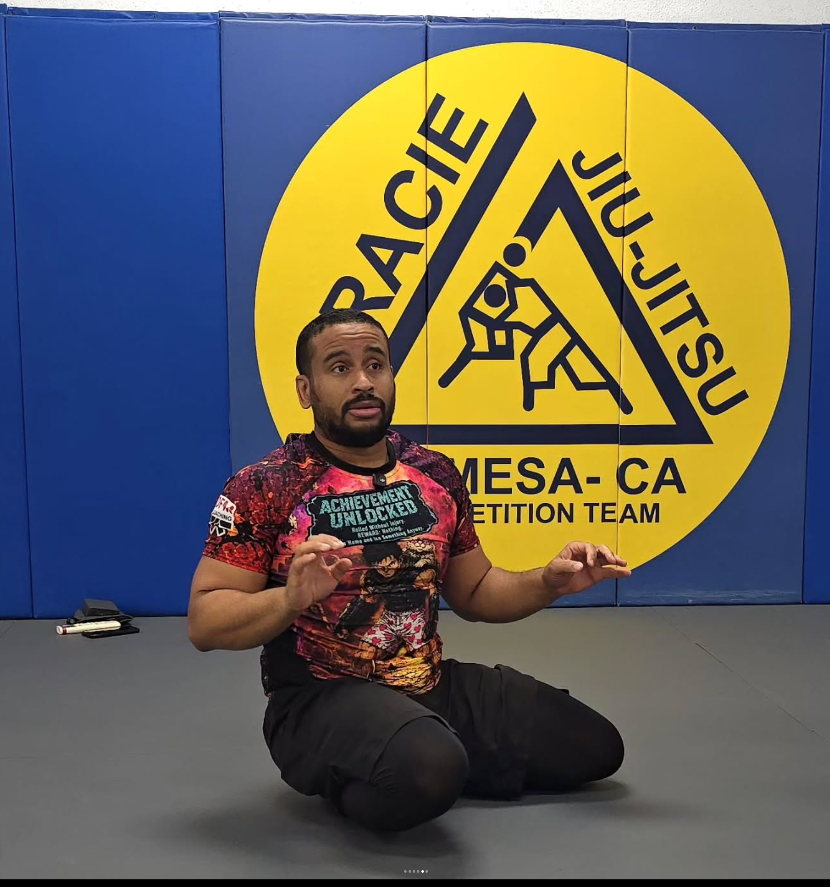 A man kneeling on a gray mat in front of a large yellow and black sign that reads "TRIACE OF JIU JITSU MESA-CALIFORNIA" and features a graphic of two people practicing jiu jitsu. The man is holding a black sign that says "ACHIEVEMENT UNLOCKED" and has a small logo on his sleeve. There is a marker and cloth on the table in front of him.
