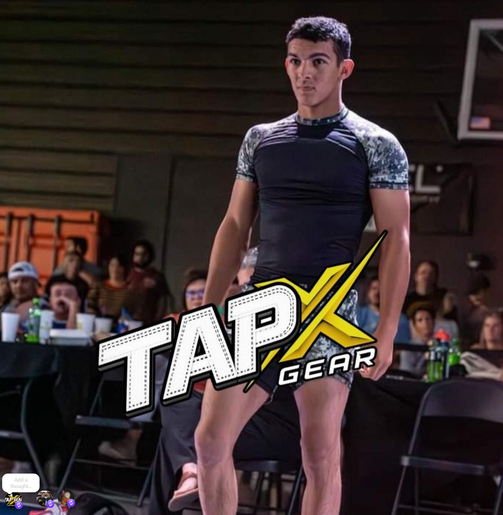 A man in black and patterned sportswear stands in a gym with a group of people sitting at tables behind him.