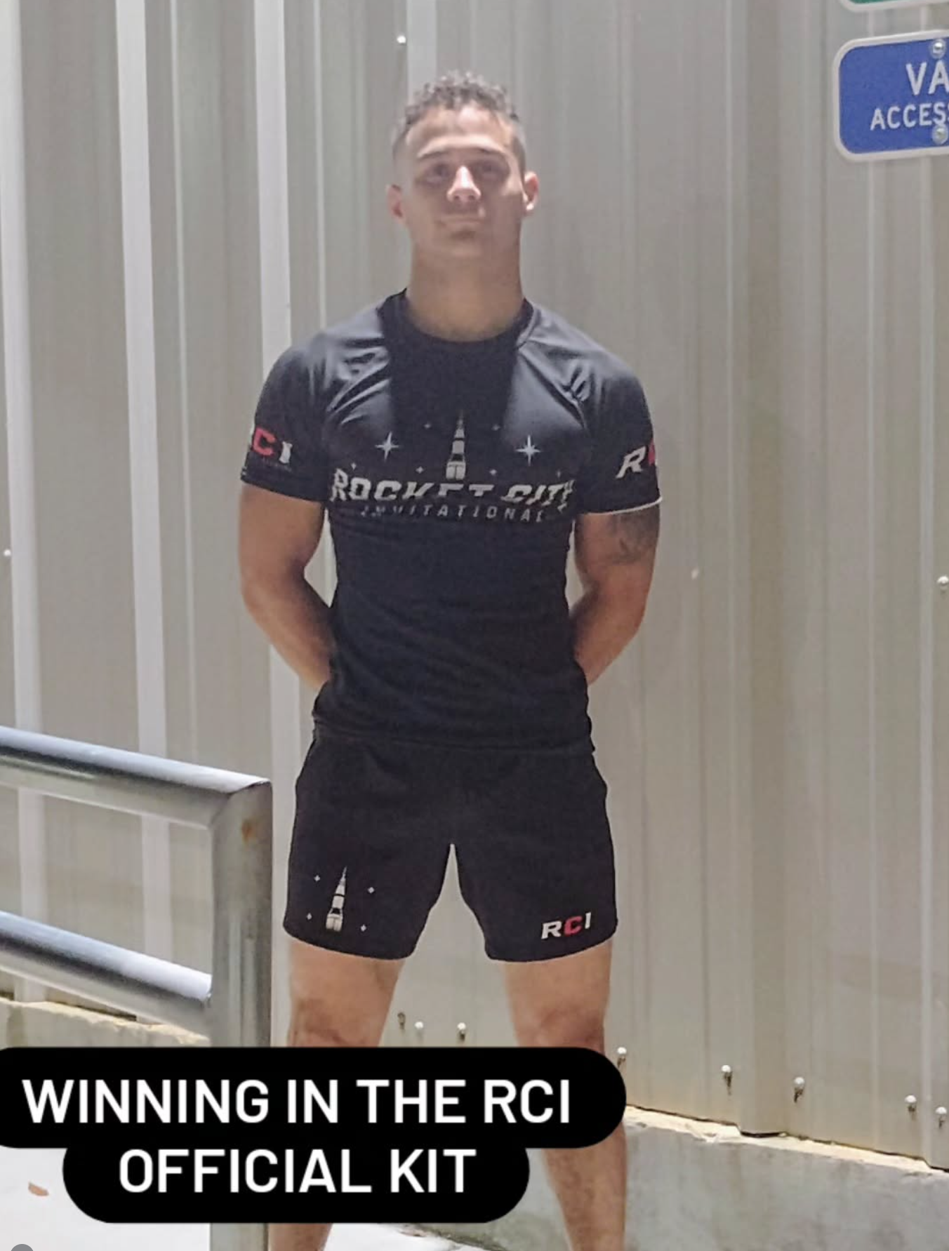 A young man wearing a black Rockt City International athletic kit standing outdoors against a metal wall, with a railing and a sign that says 'VAN ACCESS.'