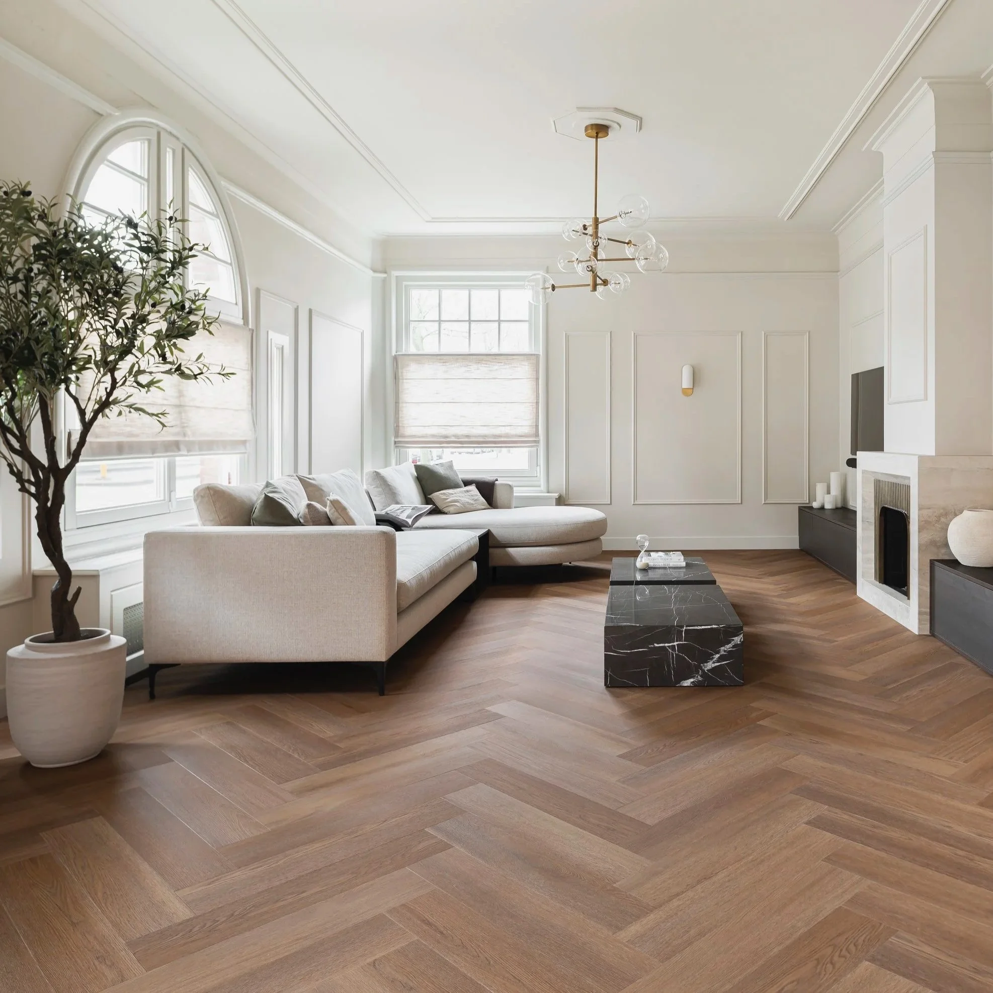 Bright and minimalist living room with white walls, large windows with beige roman shades, a beige sectional sofa with pillows, a black marble coffee table, a modern chandelier, wooden herringbone flooring, and a fireplace with decorative items.